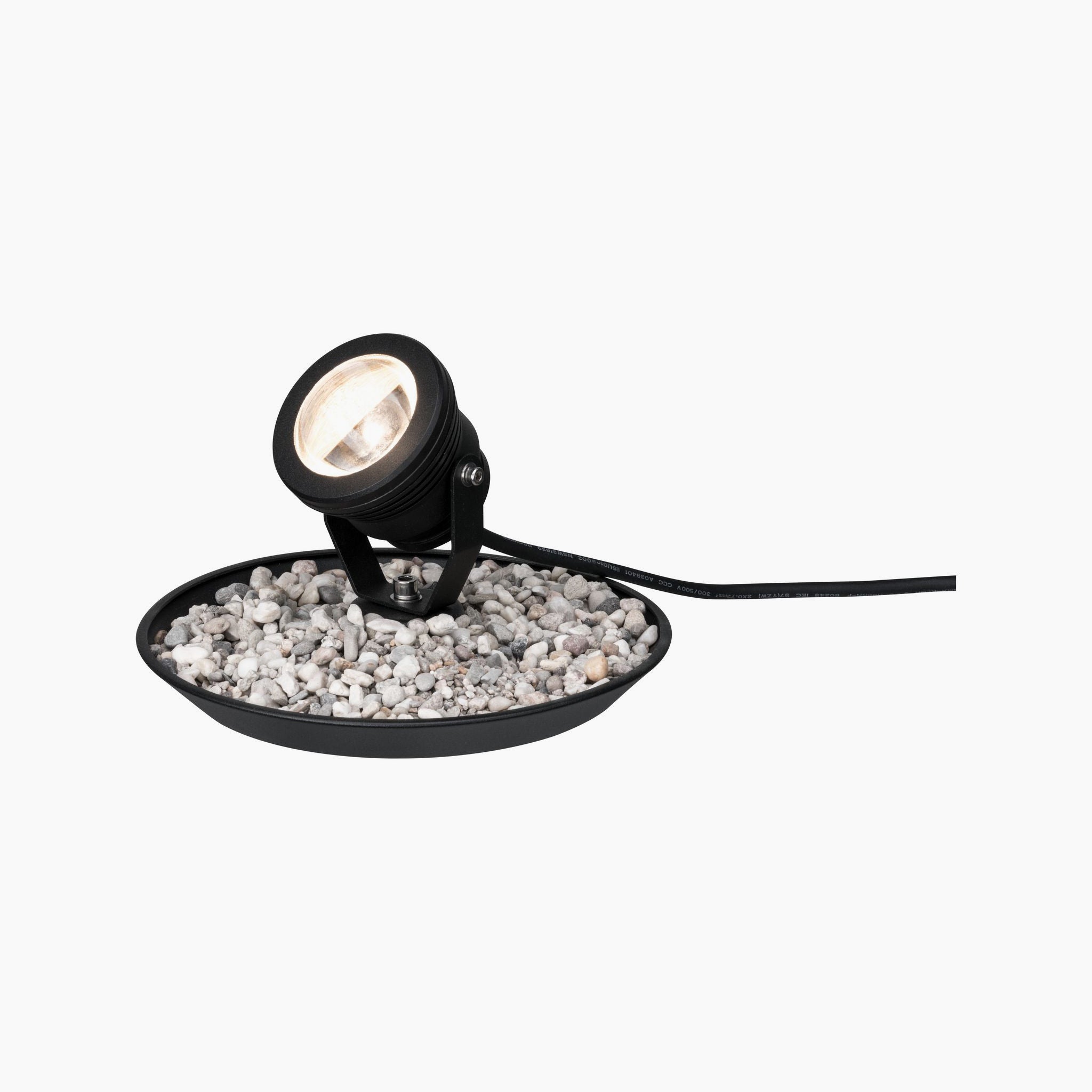 The Paulmann - Plug & Shine Outdoor 2.8W LED Pond Spotlight in black is elegantly positioned on a tray filled with small white pebbles, seamlessly connected by a cable, making it ideal for illuminating your outdoor spaces.