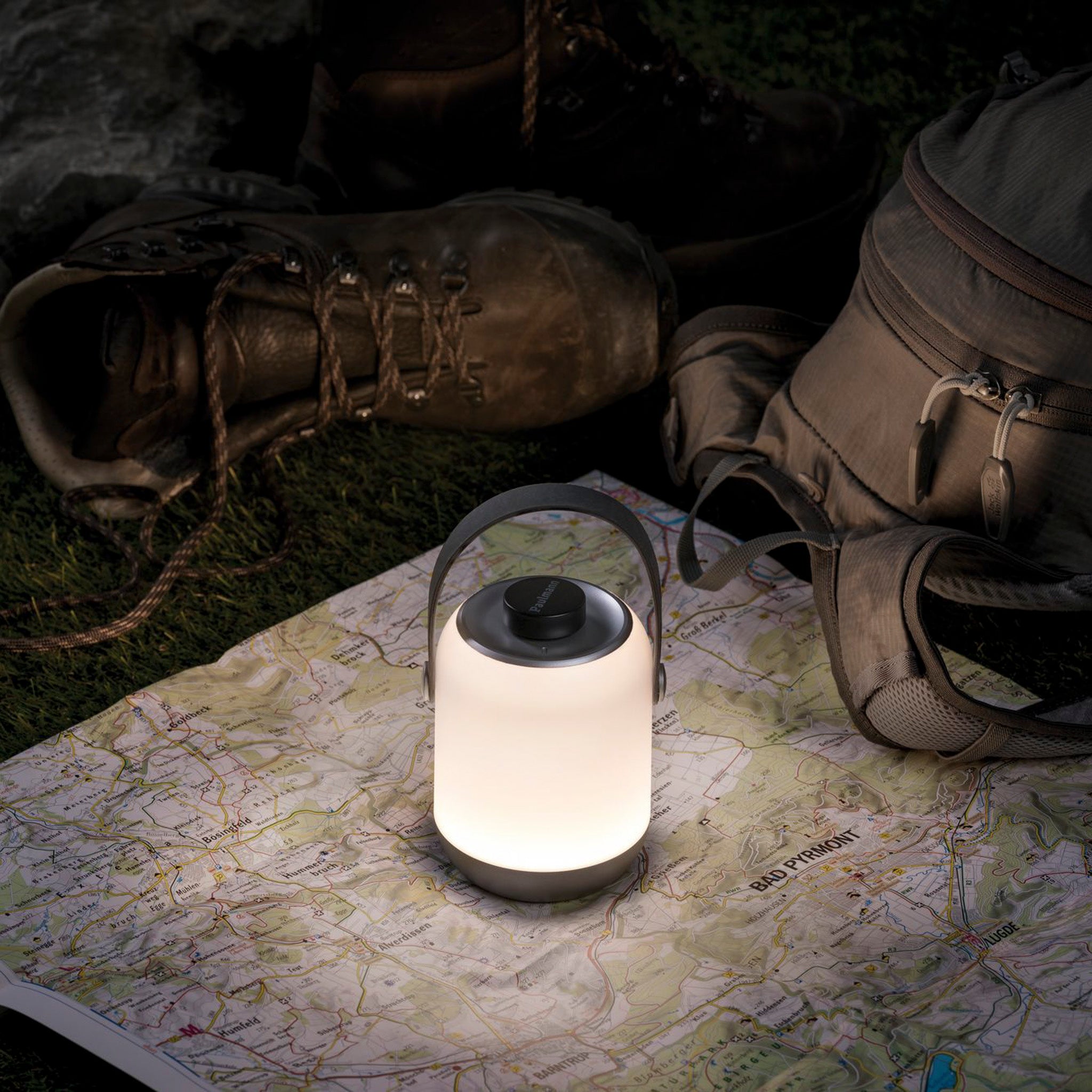 The Outdoor Clutch 6W LED Mobile Table Lamp in Grey glows on a map with hiking boots and a backpack in the background, all set on grass.