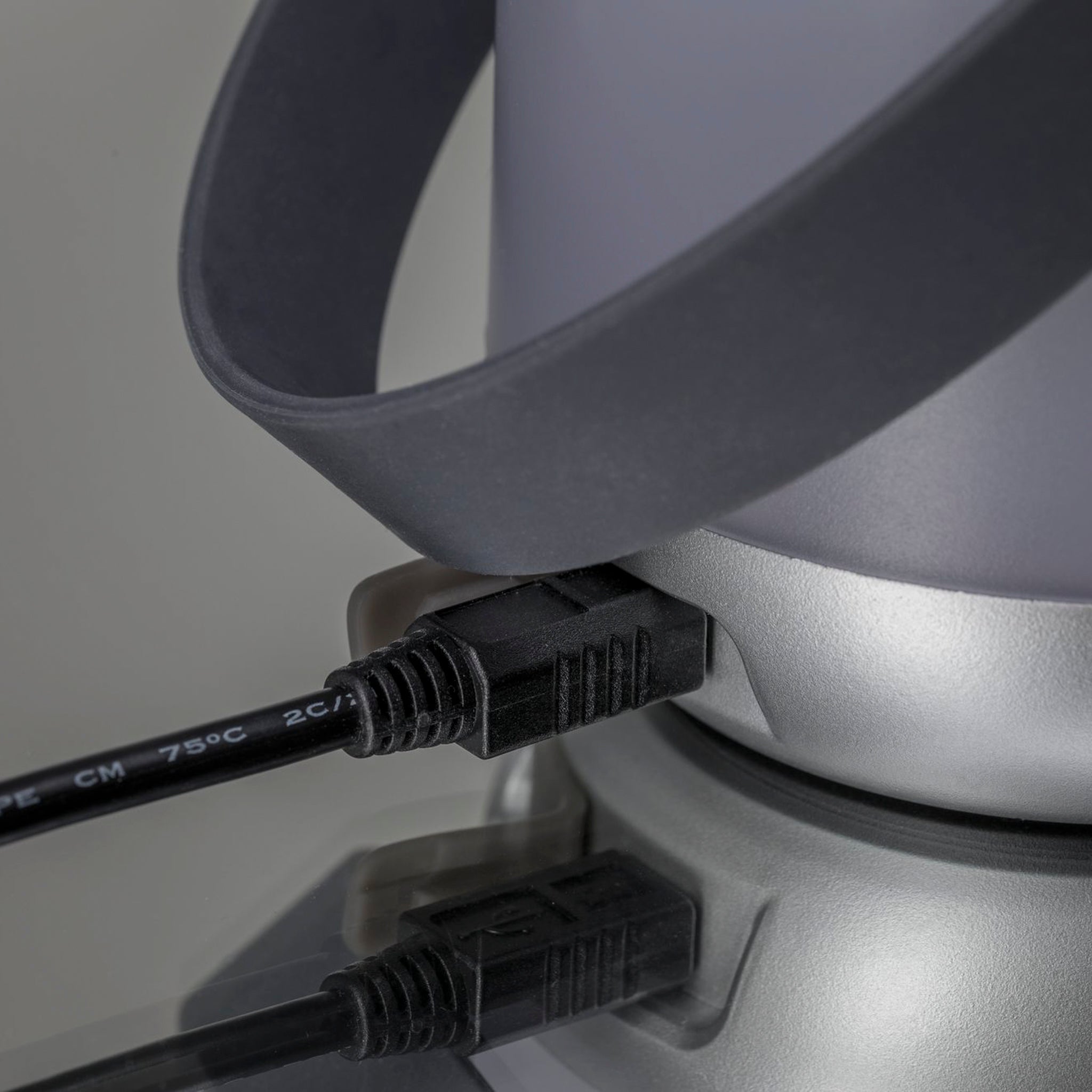 Close-up of a black cable plugged into the sleek silver Outdoor Clutch 6W LED Mobile Table Lamp in Grey, with a reflective surface underneath. The portable lamp enhances the modern aesthetic.