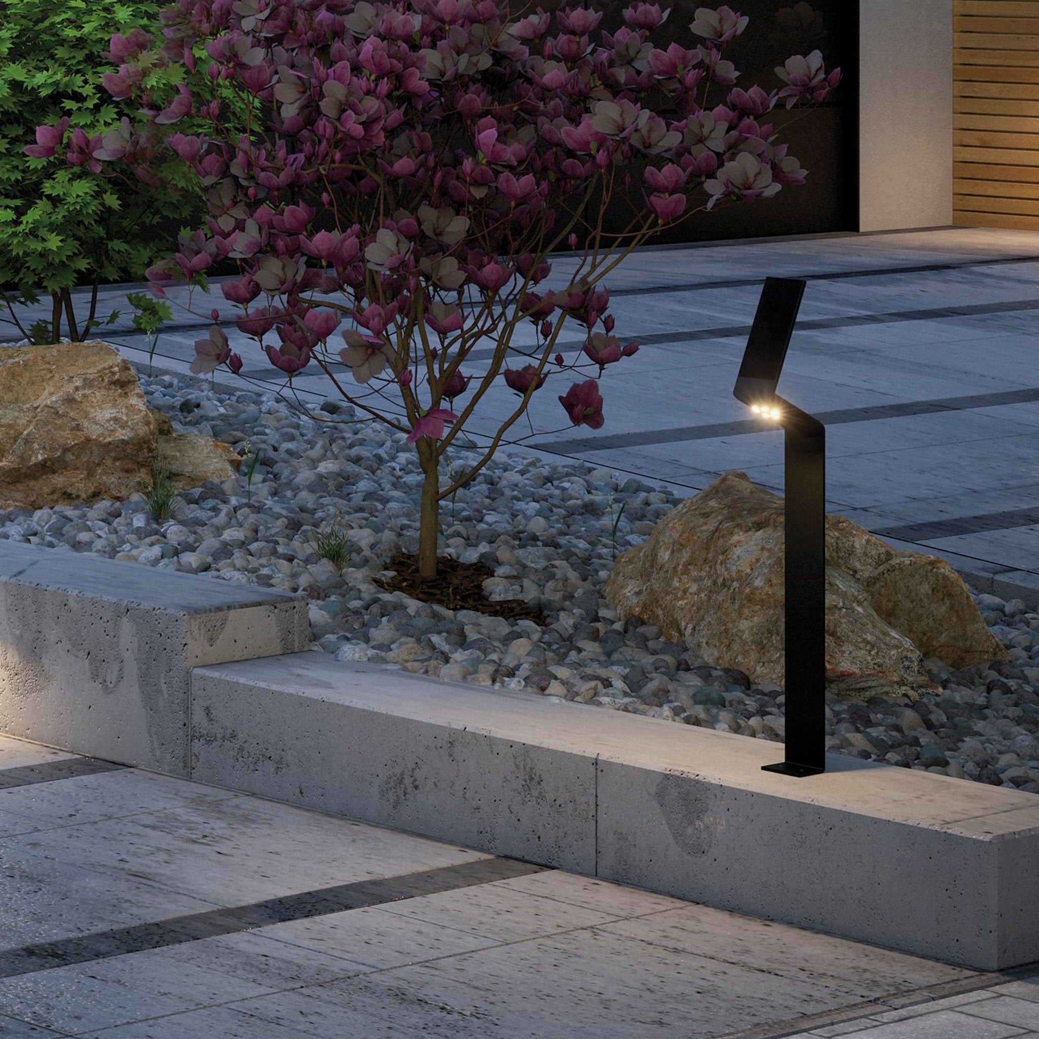 Beside a flowerbed adorned with rocks and a flowering shrub on a stone pavement at dusk stands the Paulmann Outdoor Ilias 1.2W LED Solar Bollard Light in Anthracite, featuring motion detection. Its advanced solar technology efficiently recharges the battery, ensuring illumination throughout the night.