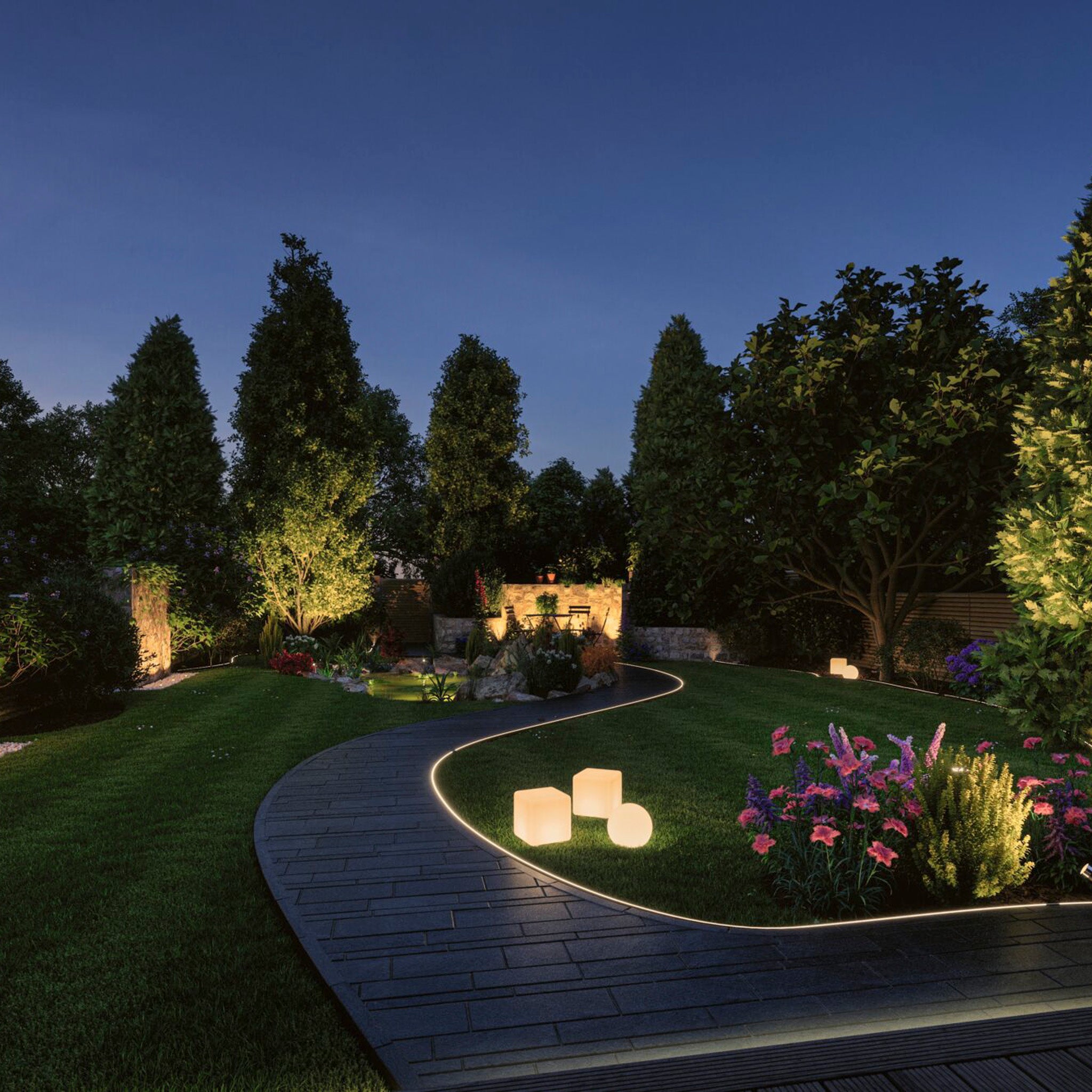 The garden path is beautifully illuminated at night, surrounded by trees, flowers, and glowing cube lights. The magic is enhanced by outdoor lighting using the Paulmann - Plug & Shine Outdoor 75VA Power Supply Transformer in White.