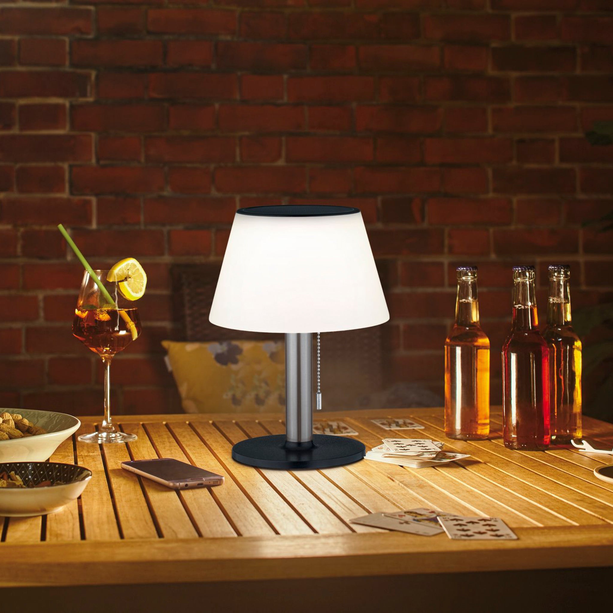 The Outdoor Lillesol 0.8W LED Solar Table Lamp with 3-Step Dim in Stainless Steel illuminates the table, casting a warm glow on the drinks, snacks, and playing cards in the cozy, brick-walled room.