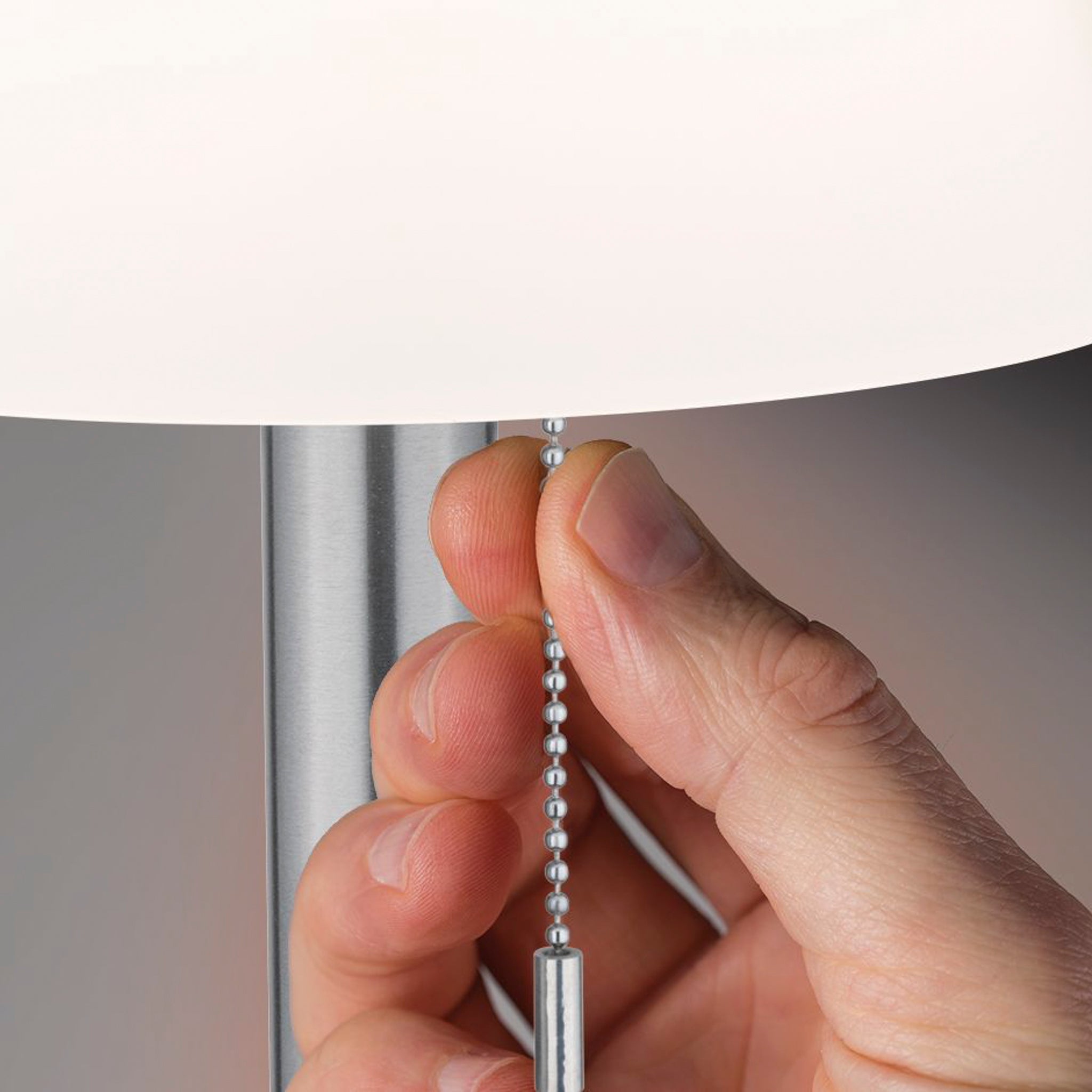 A hand pulls the chain to turn on the Outdoor Lillesol 0.8W LED Solar Table Lamp with 3-Step Dim in Stainless Steel. The lamp has a silver base and a white shade, resembling the elegance of indoor garden lighting.