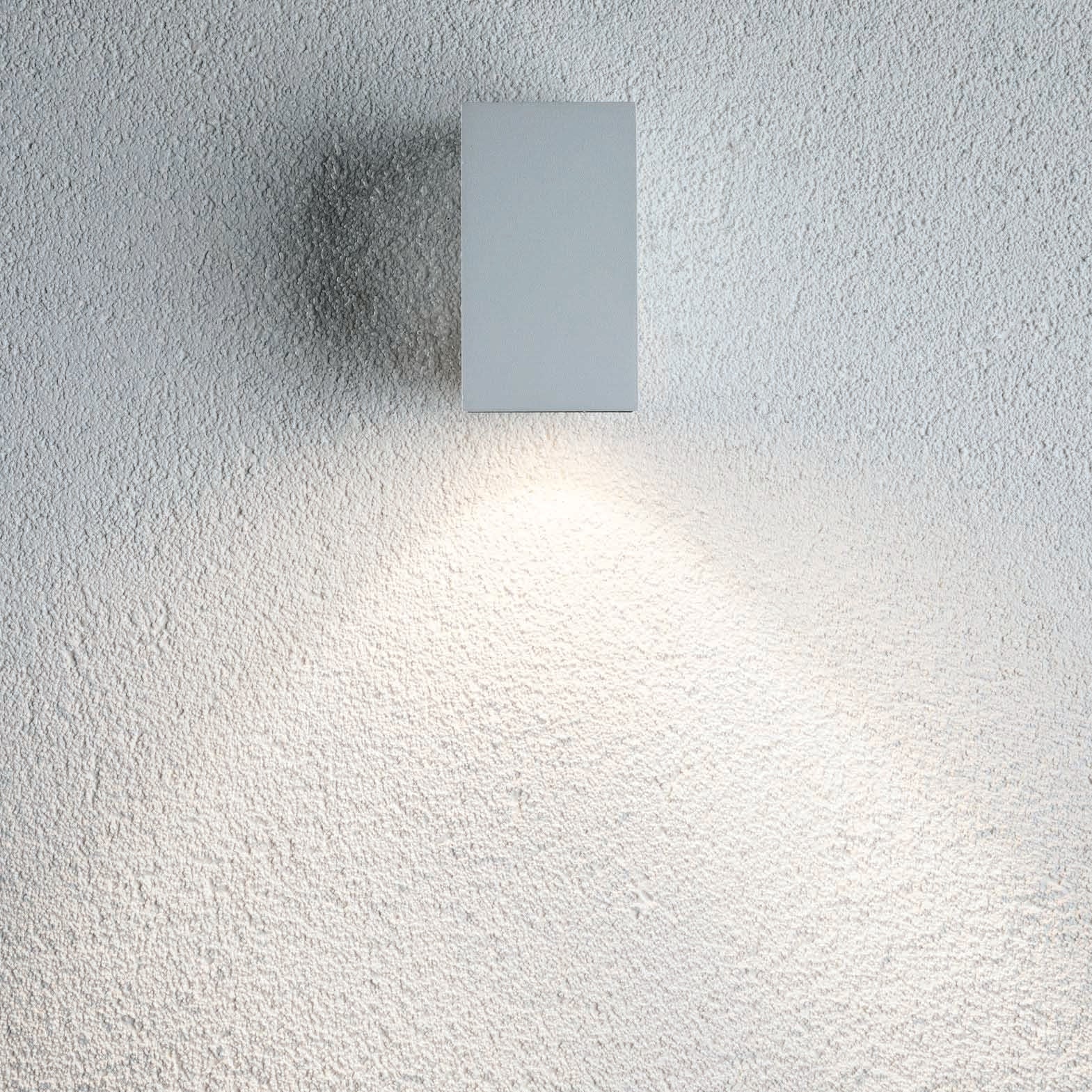 The Outdoor Flame 5.2W LED Single Output Wall Light in White is a minimalist, surface-mounted wall luminaire that emits efficient, soft light, illuminating the textured white surface of the wall below it and creating attractive lighting effects.