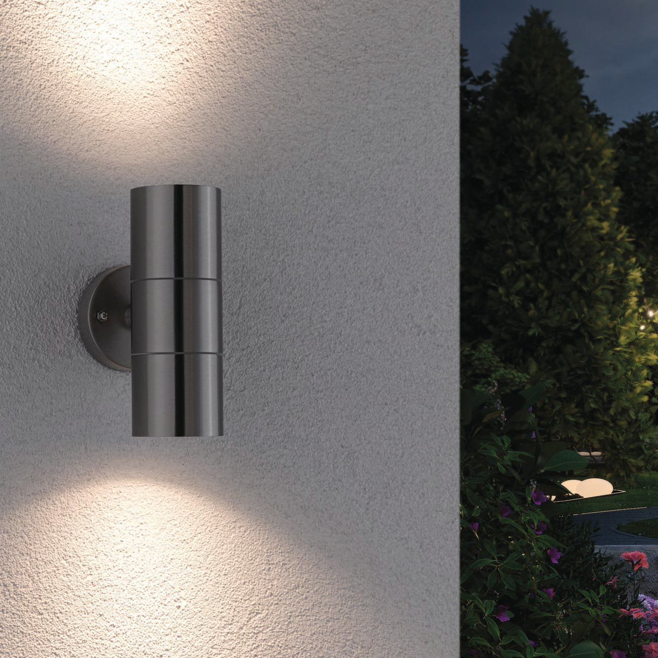 The Paulmann Outdoor Flame 10W LED Dual Output Wall Light in Stainless Steel elegantly illuminates the garden, subtly highlighting trees and plants in the background with its advanced LED technology for stunning lighting effects.