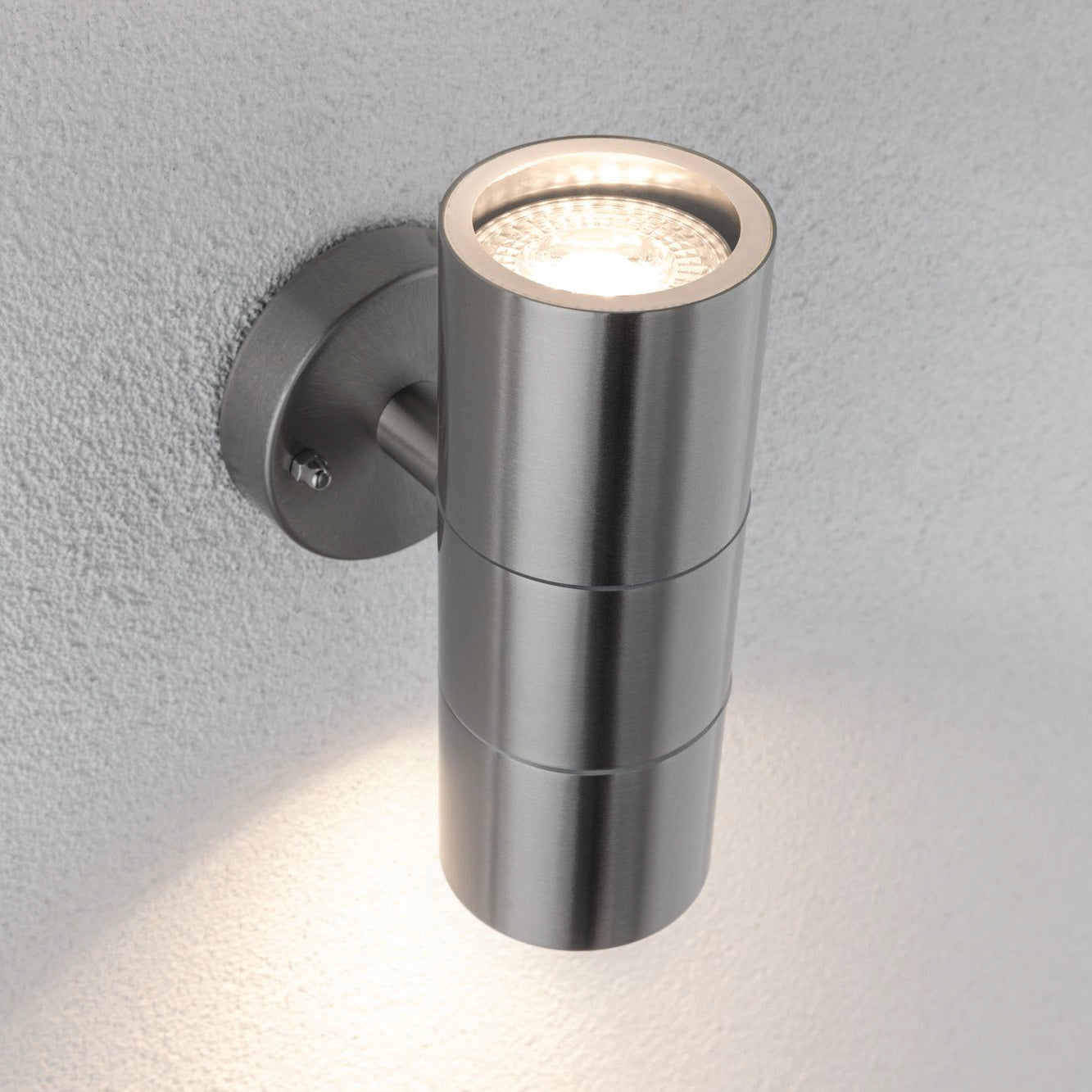 The Paulmann - Outdoor Flame 10W LED Dual Output Wall Light in Stainless Steel, a wall-mounted cylindrical luminaire, projects lighting effects both upward and downward on a white textured wall.