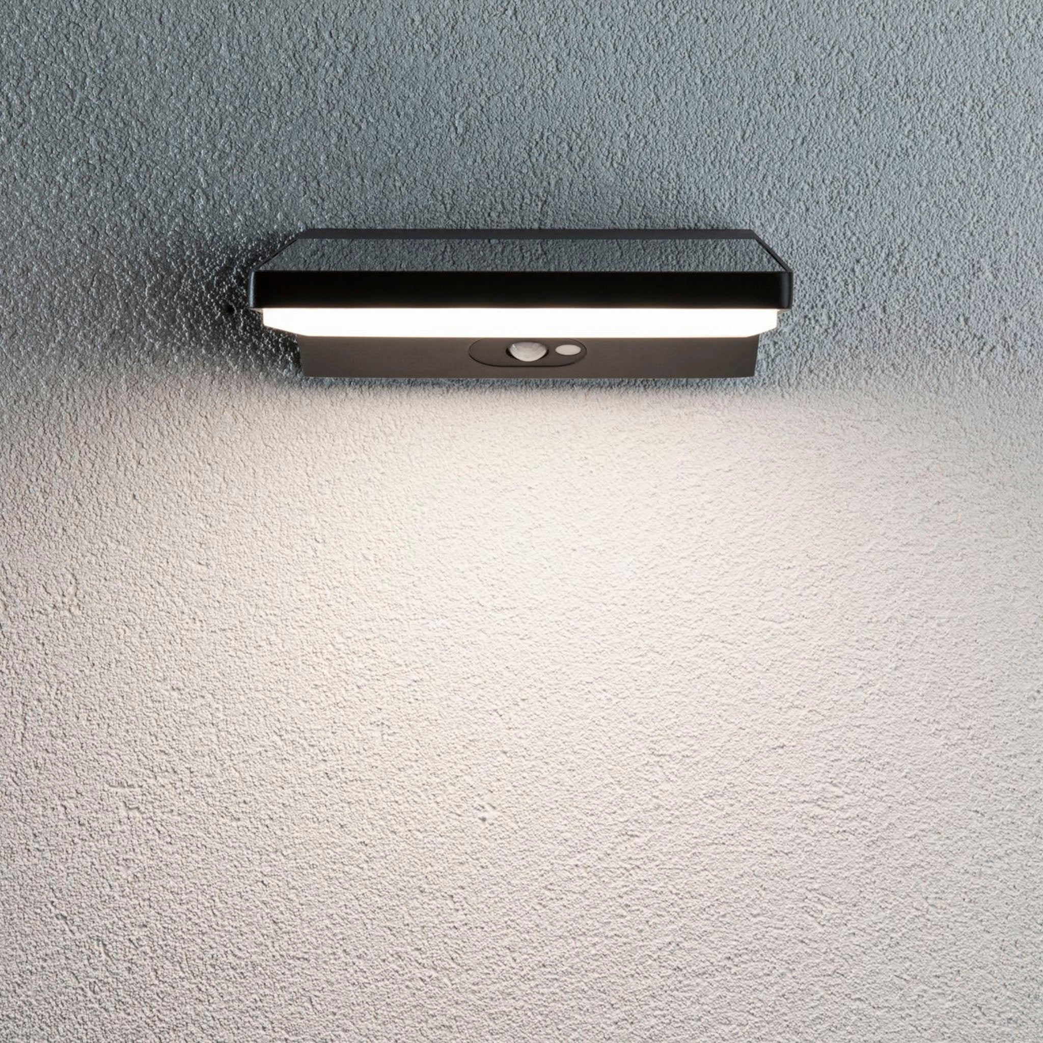 The Paulmann - Outdoor Ronas 5W LED Solar Wall Light with Motion Detection in Dark Grey illuminates a textured surface with its modern, rectangular design.