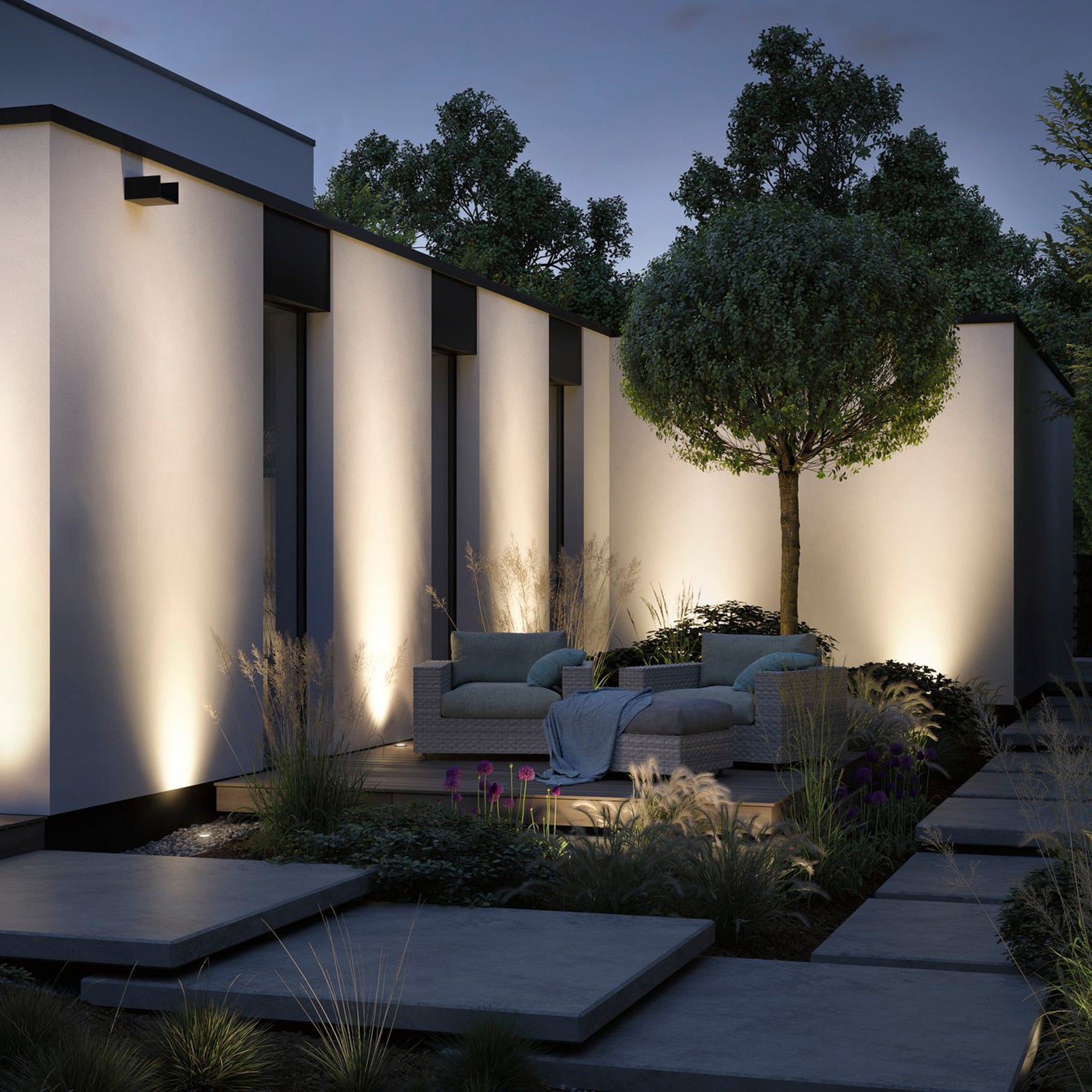 A modern outdoor patio featuring cozy seating and the Paulmann Plug & Shine Outdoor 11W LED Recessed Floor 8.2cm Spotlight in Grey provides ambient lighting, while surrounding trees enhance the serene evening atmosphere.