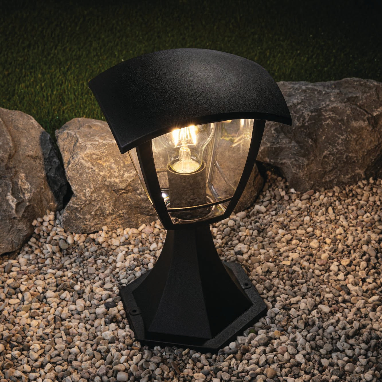 The Paulmann Outdoor Classic Curved 12W LED Bollard Light in Black enhances the enchanting scene on a gravel pathway, casting illumination against a backdrop of rocks and grass.