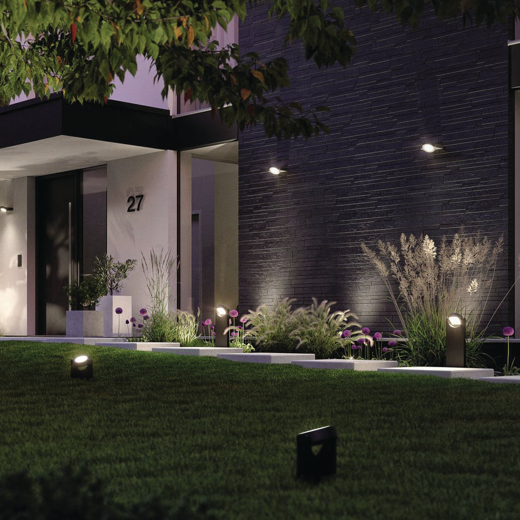 The modern building entrance at night is enchanting, with exterior lighting accentuating the illuminated pathway. The Paulmann Outdoor Swivea 8W LED Bollard Lights in anthracite guide the way through lush greenery and vibrant purple flowers, creating a captivating ambiance.