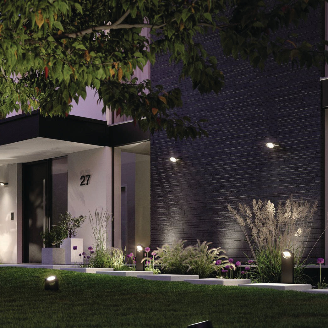 A modern house entrance featuring a dark stone wall, Paulmann's Outdoor Swivea 8W LED Garden Spotlights in Anthracite casting warm white light, and lush greenery, all highlighting contemporary design.