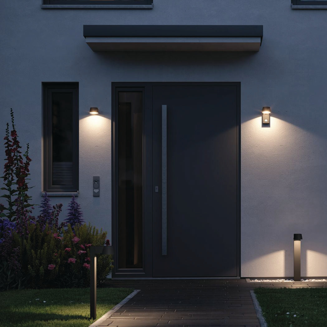 A modern entrance features a dark door illuminated by the Paulmann - Outdoor Capea 8W LED Wall Light in Anthracite, surrounded by lush plants and vibrant flowers, enhancing the appeal of the exterior walls.