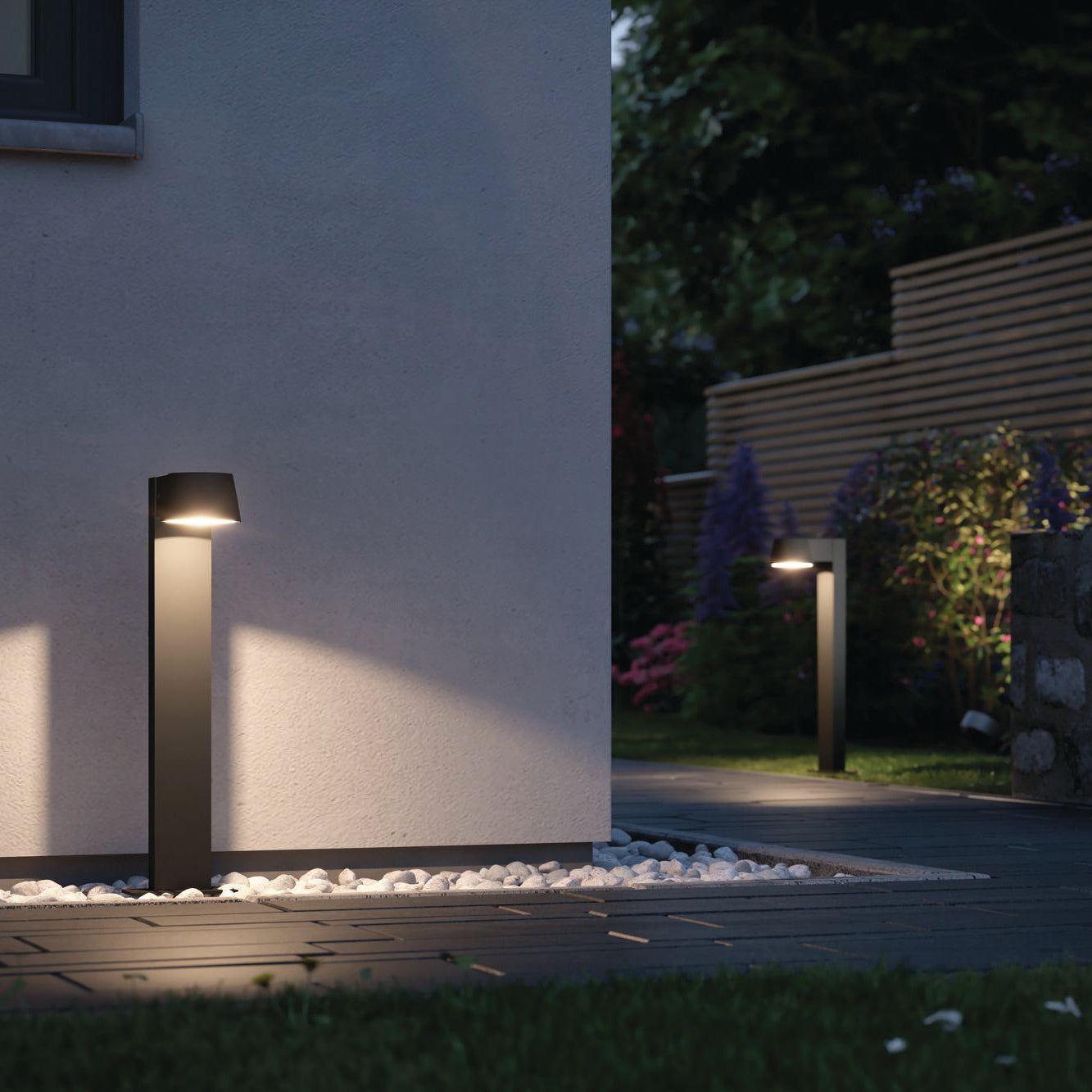 The garden path next to the white wall and greenery is illuminated by the Paulmann Outdoor Capea 8W LED Bollard Light in Anthracite. Designed with safety in mind, these waterproof bollard lights guarantee durability in all weather conditions.