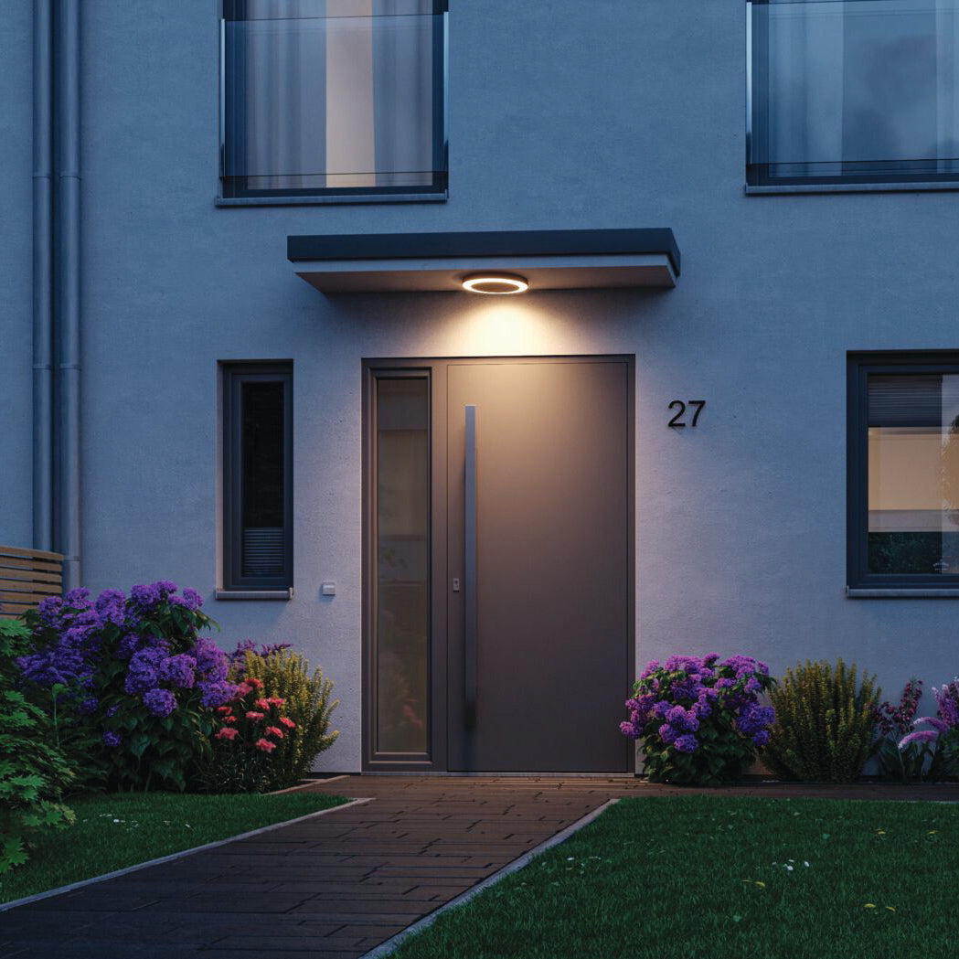 A contemporary front door with a sleek design, framed by purple flowers and greenery, is lit by the Paulmann - Outdoor Circula Dusk 15W LED Smart Home Zigbee 3.0 Warm Ceiling Light in Anthracite, which features motion detection.