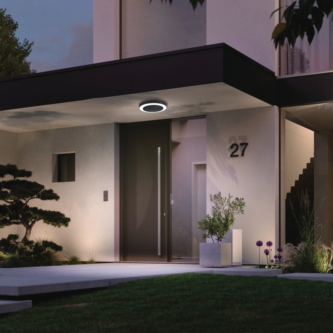 As evening falls, the entrance of the contemporary house is tastefully lit by a Paulmann Outdoor Circula Dusk 15W LED Ceiling Light in Anthracite. The gentle glow accentuates the surrounding lush plants, while the built-in motion detection feature ensures effortless illumination with house number 27 prominently displayed.