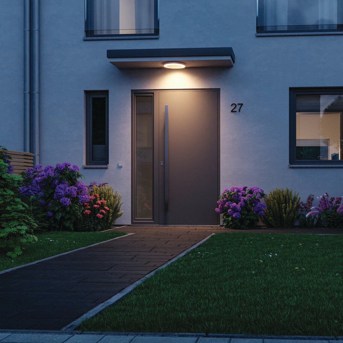 A contemporary building entrance at night showcases an illuminated door enhanced by the Paulmann - Outdoor Circula Dusk 15W LED Smart Home Zigbee 3.0 Warm Ceiling Light with Motion Detection in White. The pathway, lined with greenery and purple flowers, gains from the Tunable Warm function, creating a cozy ambiance.