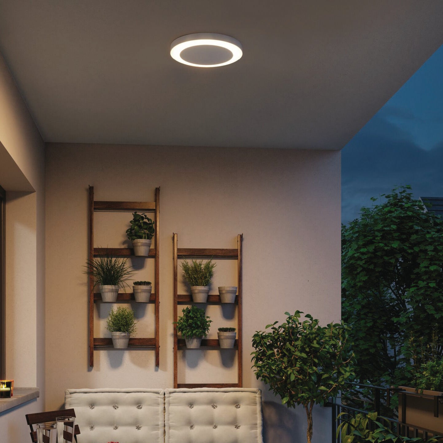 Cozy balcony with potted plants on wooden shelves and the Paulmann Outdoor Circula Dusk 15W LED Ceiling Light in white, featuring motion detection, casting a warm glow above the cushioned seating.