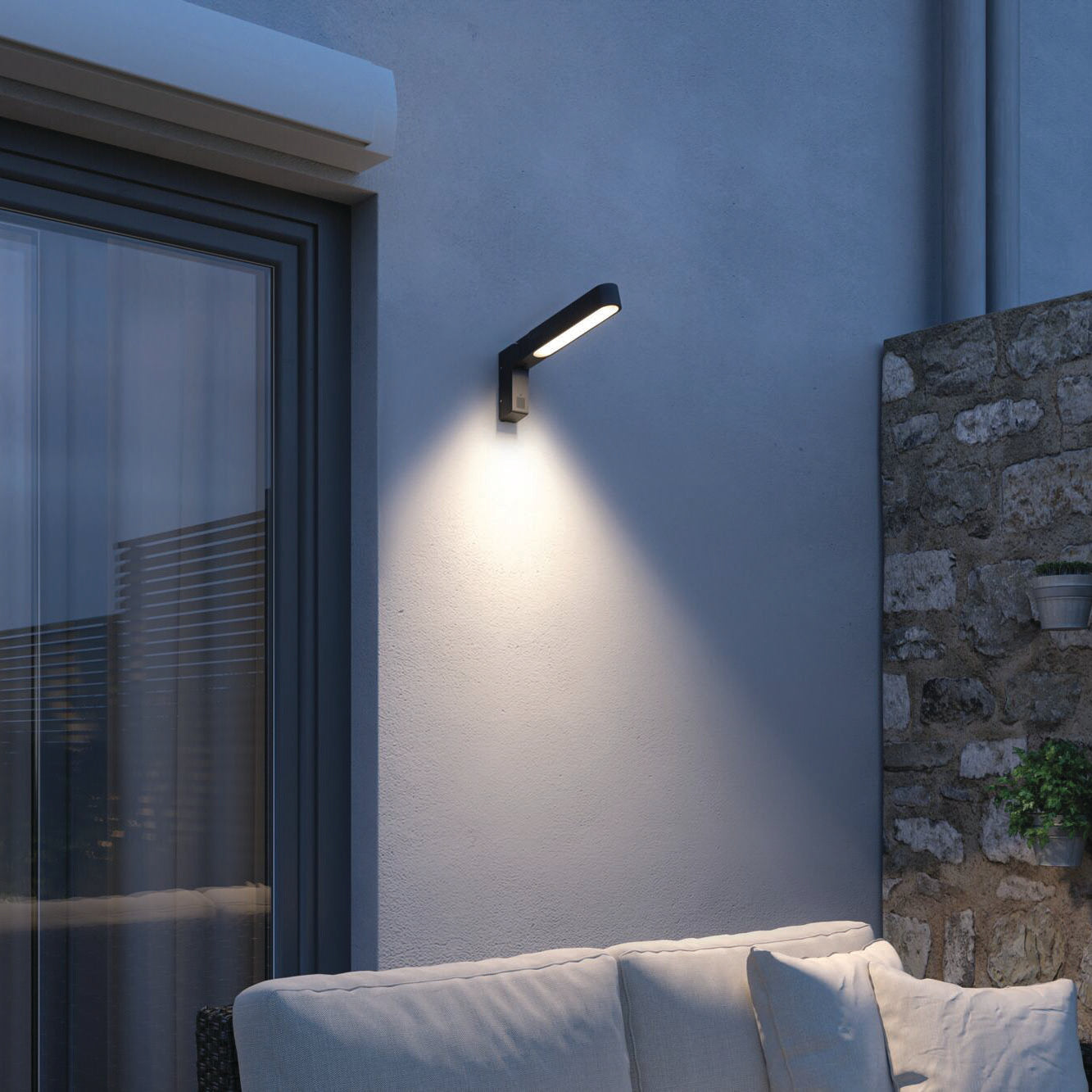 The Paulmann - Outdoor Ito 7.5W LED Vertical Wall Light with Motion Detection in Anthracite enhances the ambiance of a modern patio, elegantly illuminating the stone wall and sofa, while providing IP44 splash protection.