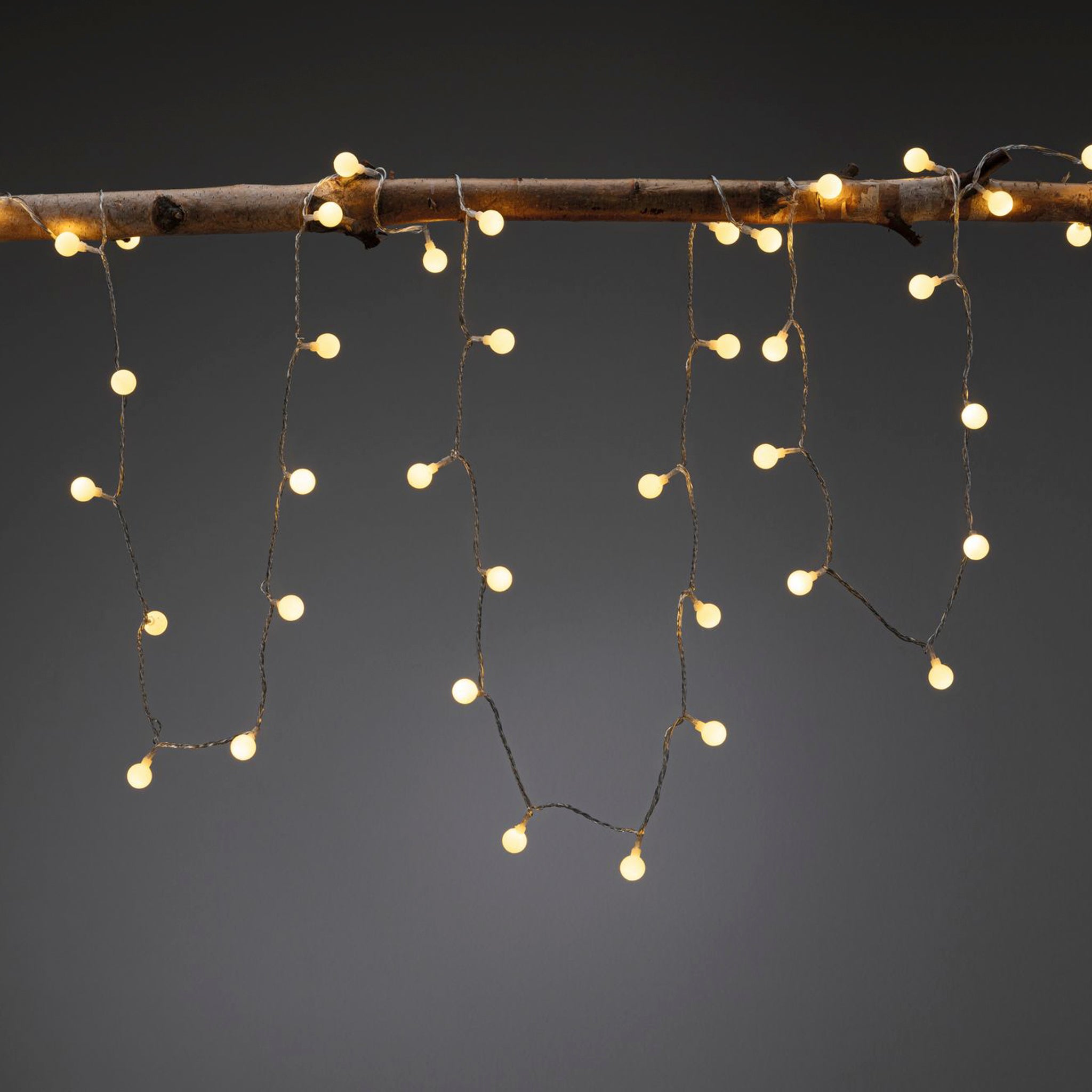 The Paulmann Plug & Shine Outdoor 1.8W LED 5m warm white fairy lights, set against a dark background and suspended from a wooden rod, create an enchanting outdoor lighting display.