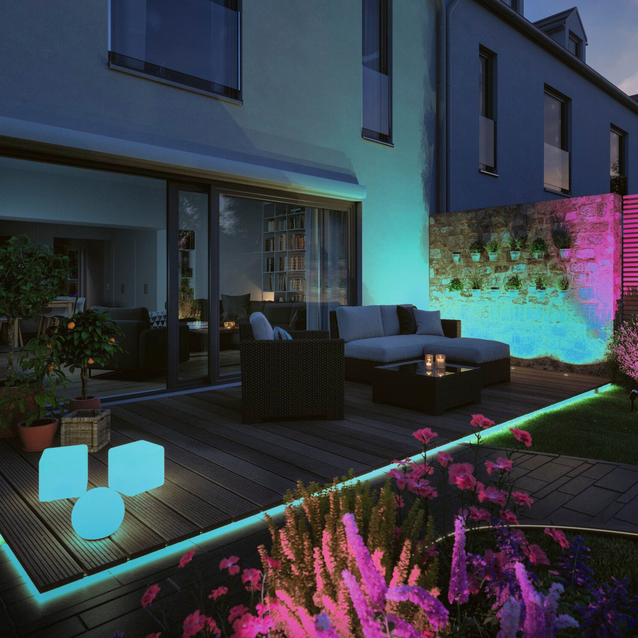 A contemporary patio featuring glowing furniture and plants is illuminated by the Paulmann Plug & Shine Outdoor 11W LED 2m Smooth Strip with Smart Home Zigbee 3.0 in RGBW, casting blue and pink hues against a stone wall at night.