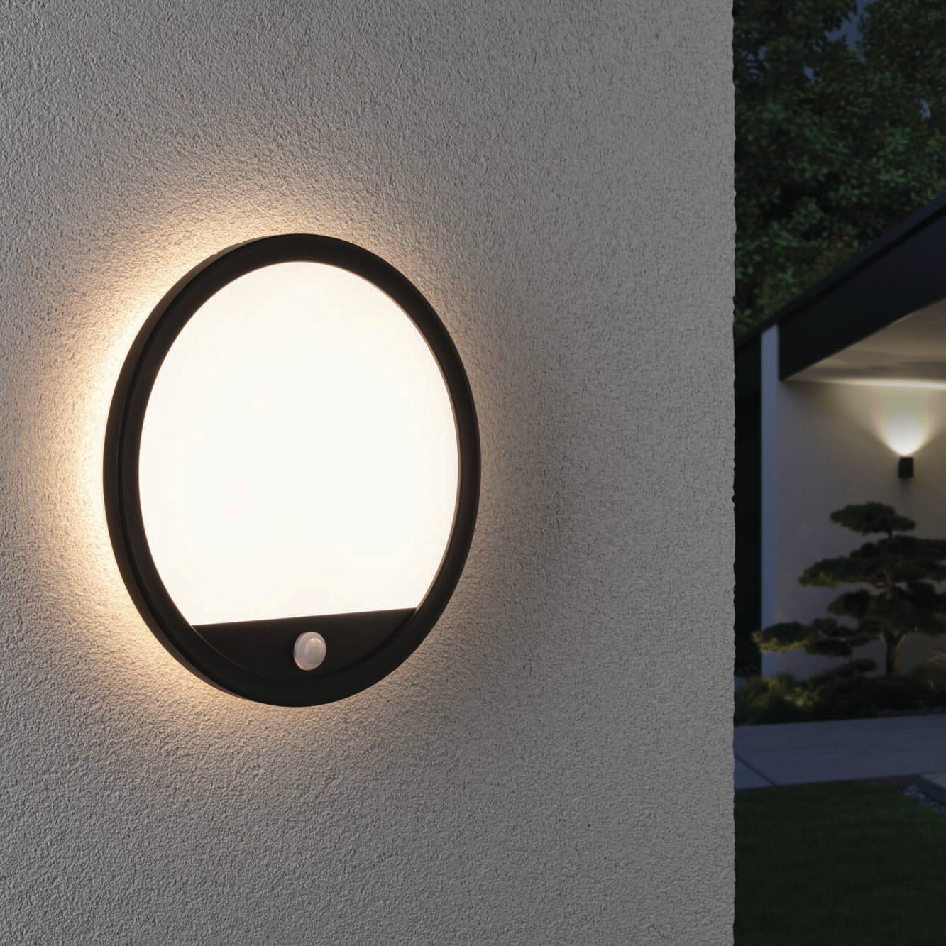 The Outdoor Lamina 15.5W LED Backlit Round Wall Light with Motion Detection in Black is illuminated on the side of a building at dusk, casting a cozy warm white light. These stylish outdoor luminaires enhance the ambiance and provide functional elegance to your exterior spaces.