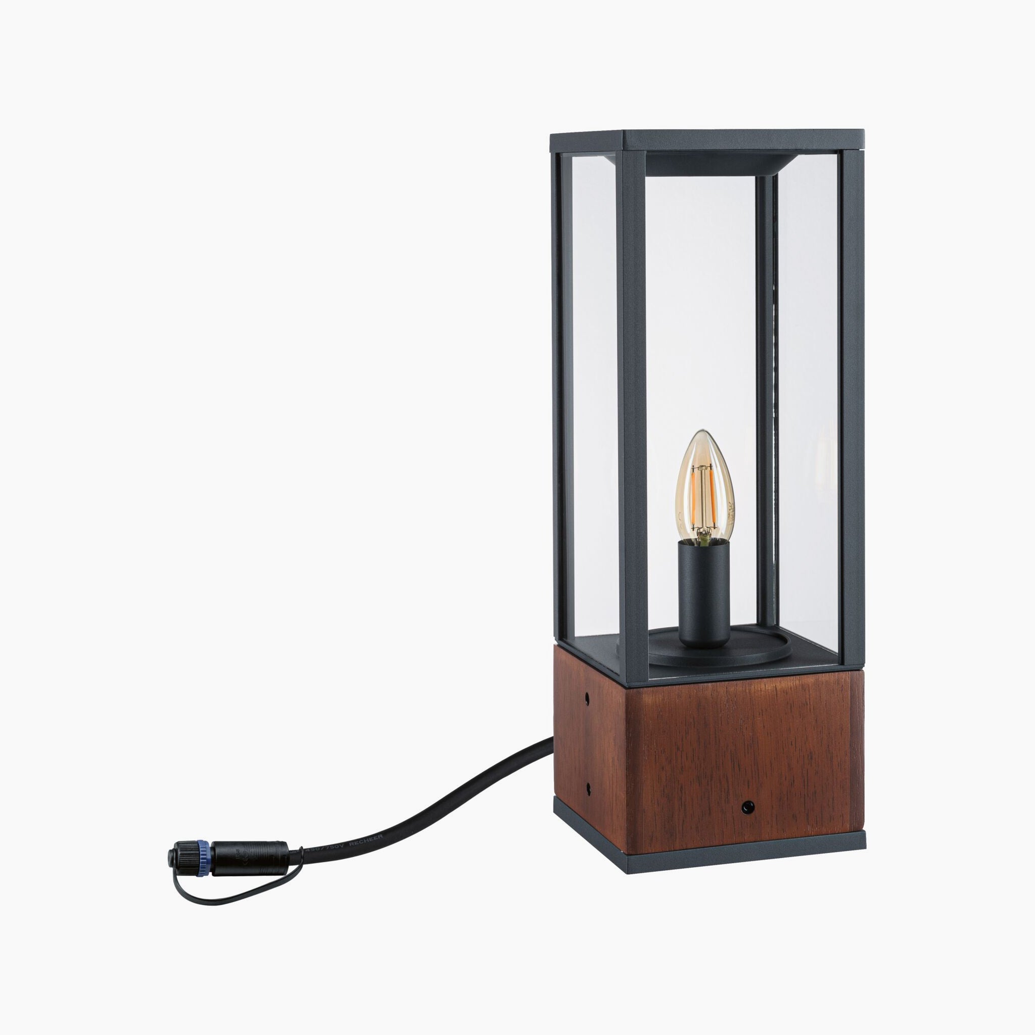 The Paulmann Plug & Shine Outdoor Vena 2W LED Wooden Single Bollard Light is a 40cm tall lantern-style lighting fixture featuring a wooden base, glass sides, and an exposed bulb connected by a black cord, offering atmospheric lighting ideal for outdoor spaces.