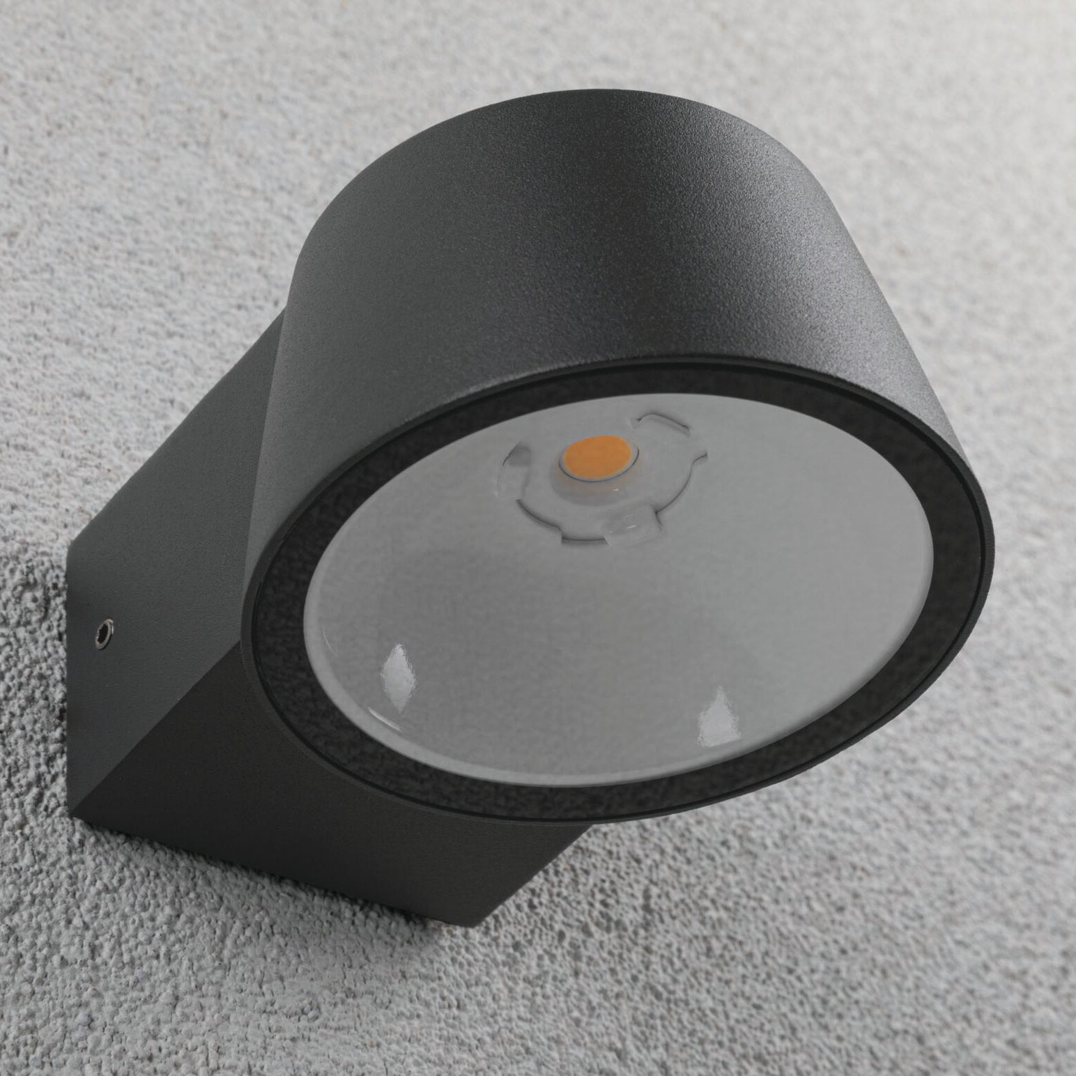 An anthracite, wall-mounted Outdoor Capea 8W LED Wall Light with an insect-friendly feature and a circular design, casting a warm gold light against a light grey textured wall.