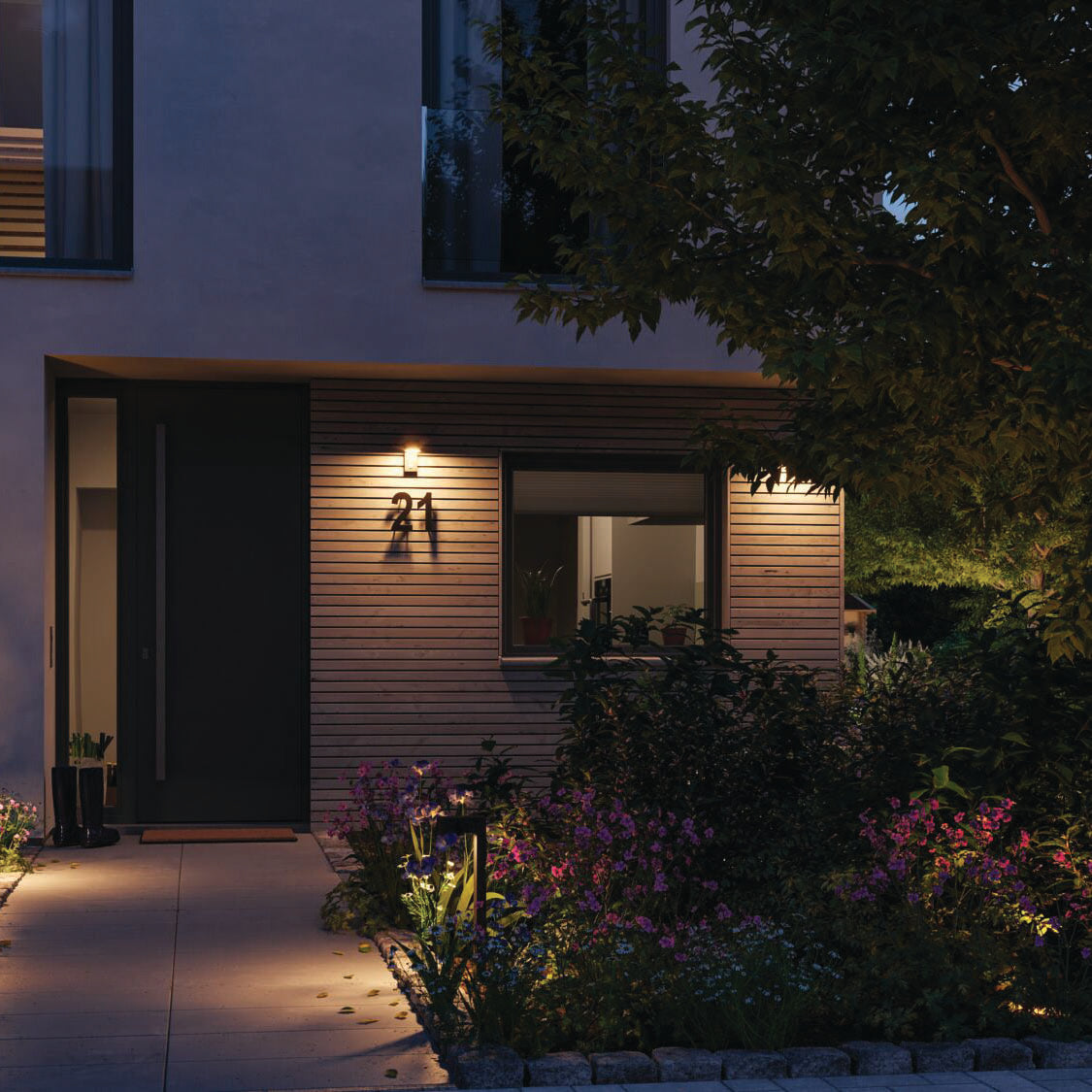 A modern house entrance at dusk with a warm gold light emanating from the Outdoor Capea 8W LED Wall Light with Insect Friendly in Anthracite, surrounded by a garden of colorful, insect-friendly flowers and plants.