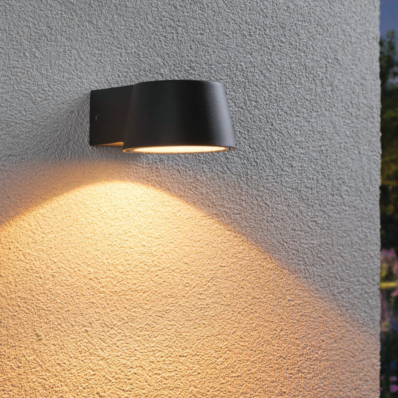 An anthracite Outdoor Capea 8W LED Wall Light with Insect Friendly casts warm gold light on a textured white exterior wall during the evening, creating an inviting atmosphere.