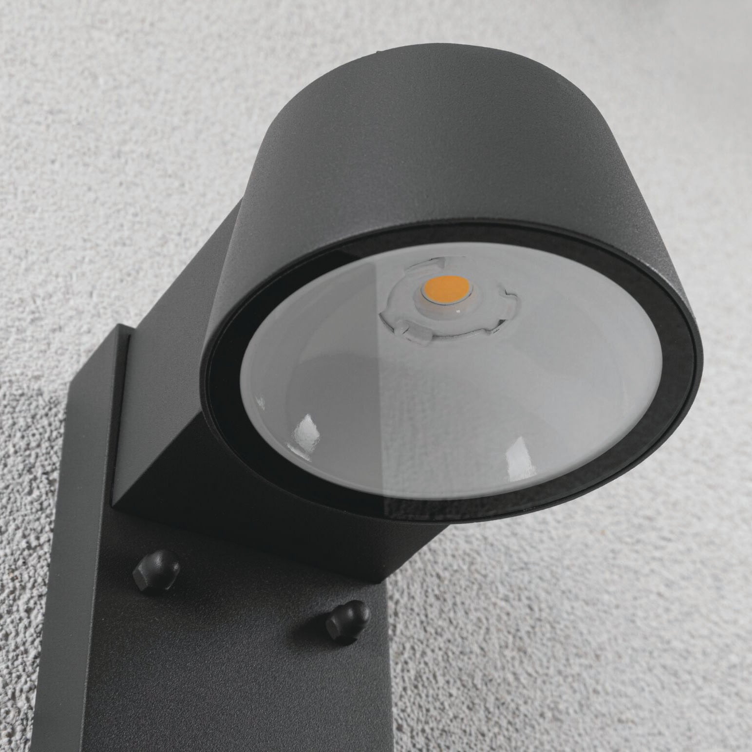 A close-up view of the cylindrical, anthracite Outdoor Capea 8W LED Wall Light featuring motion detection and insect-friendly lumination, showcasing its bulb inside, mounted on a textured wall.