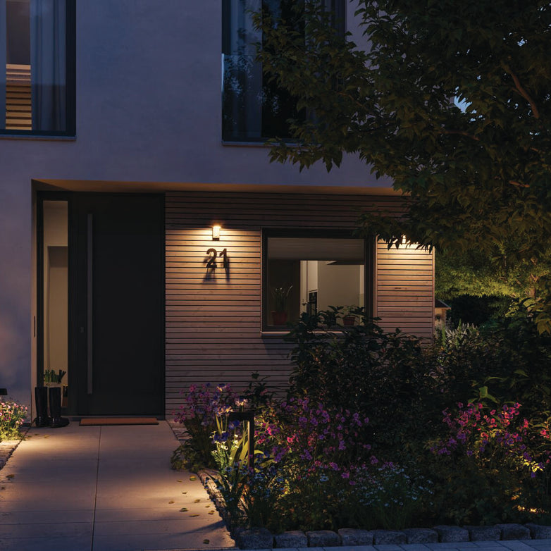 A modern home exterior at dusk features an illuminated entryway with Outdoor Capea 8W LED Wall Lights with Motion Detection and Insect Friendly Lumination in Anthracite, complemented by wooden paneling and a well-maintained, insect-friendly garden filled with plants and flowers.