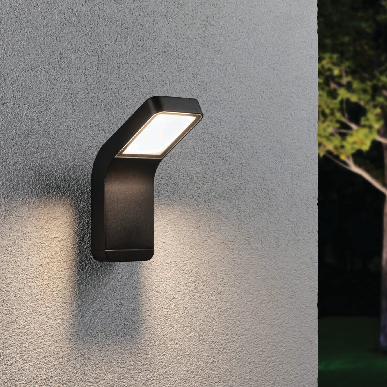 The Outdoor Kulma 9W LED Wall Light in Anthracite expertly illuminates a textured light-gray wall, with a tree visible in the background. Designed to withstand harsh coastal environments, this corrosion-resistant lighting solution provides both durability and elegance.