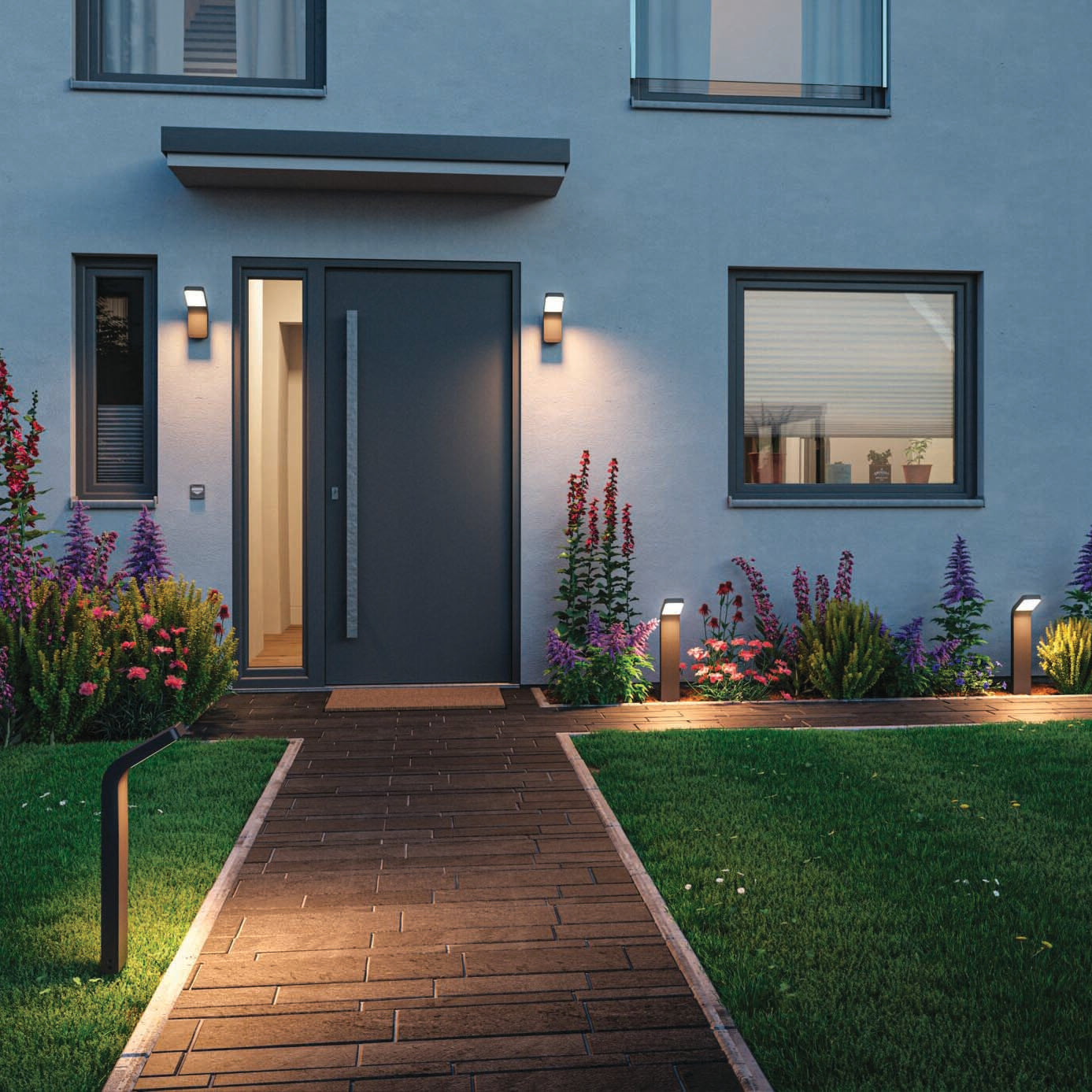 Modern house entrance with a wide door, an illuminated path featuring the Outdoor Kulma 9W LED Wall Light in Anthracite, and a garden filled with colorful flowers and green grass.