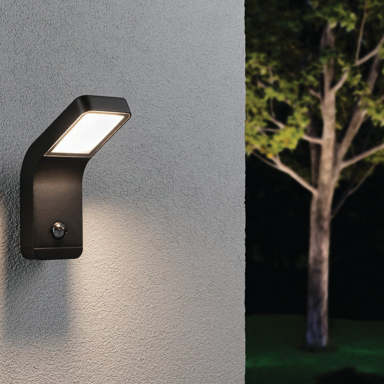 The Outdoor Kulma 9W LED Wall Light with Motion Detection in Anthracite illuminates a gray wall, creating a striking contrast with the tree and dark foliage in the background.