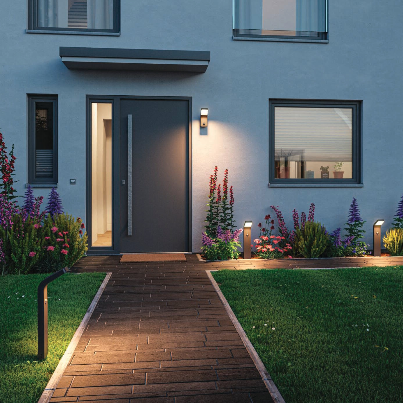 A modern house entrance with an anthracite door, pathway, garden lights featuring the Outdoor Kulma 9W LED Wall Light with Motion Detection in Anthracite, and a well-manicured lawn with colorful flowers.