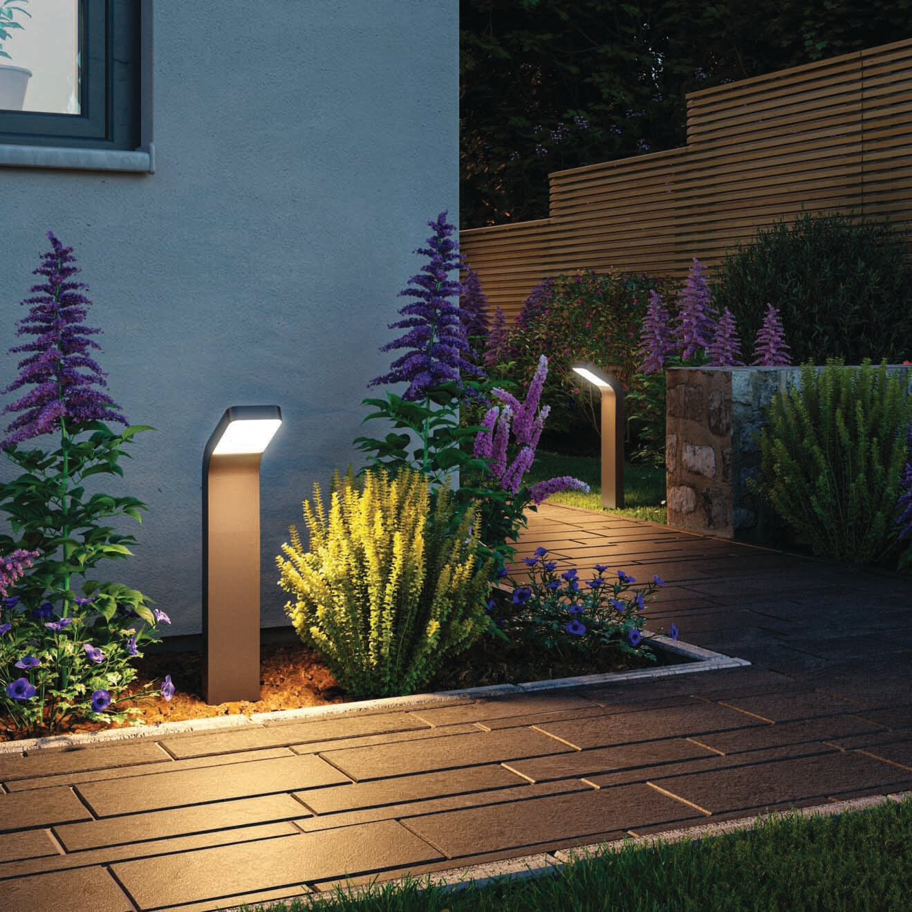 The Outdoor Kulma 9W LED Bollard Light in Anthracite illuminates a garden pathway lined with purple and yellow flowers during the evening, offering glare-free lighting.