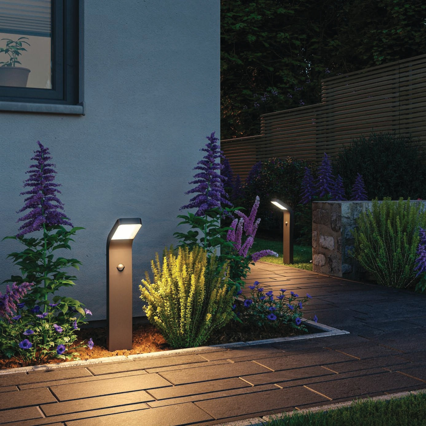 The garden path glows at night with modern Outdoor Kulma 9W LED Bollard Lights featuring motion detection and an anthracite finish, surrounded by blooming flowers and plants.