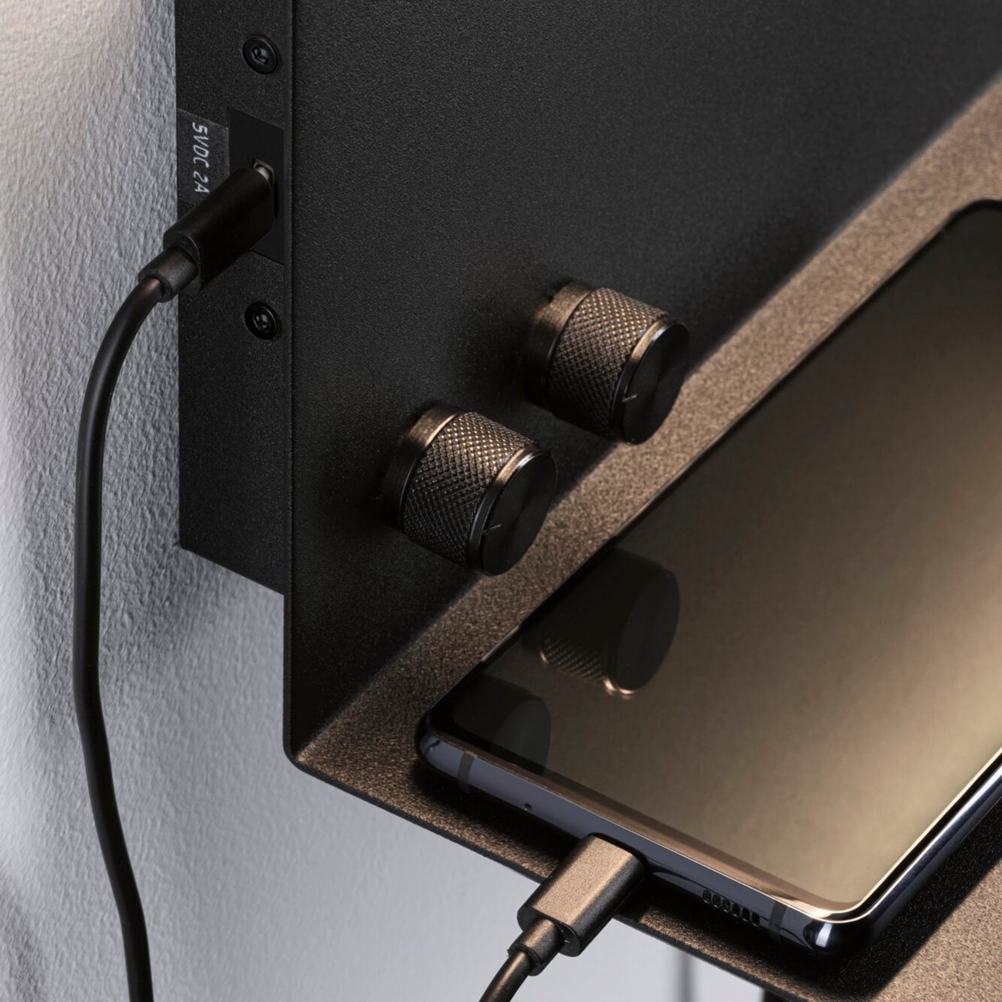 A smartphone charging on the Paulmann Jarina 7.5W LED dimmable wall light in black matt, equipped with two knobs and a handy USB port for modern devices.