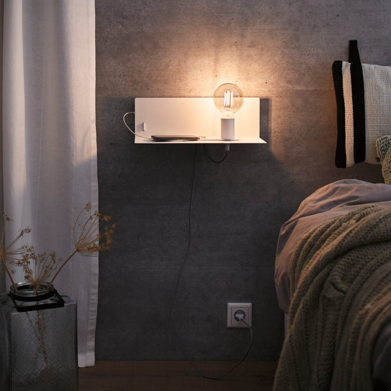 A cozy bedroom features an integrated shelf with a Devara 40W Wall Mounted Lamp with USB Port in White Matt and a lit exposed bulb lamp, next to a bed with plush bedding.