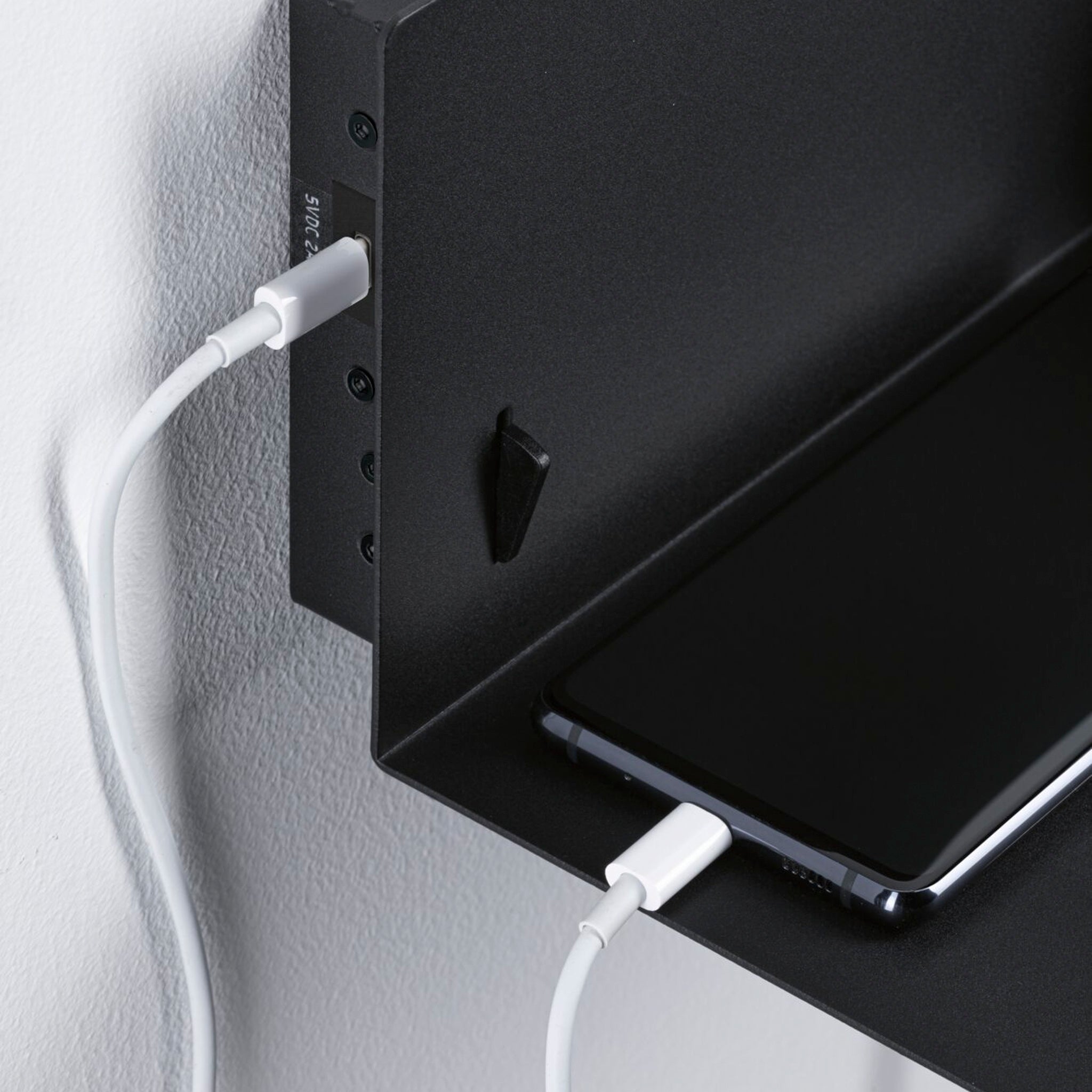 The Devara 40W Wall Mounted Lamp with USB Port in Black Matt comes with a built-in shelf and charging station, where a phone is charging alongside two white USB-C charging cables.