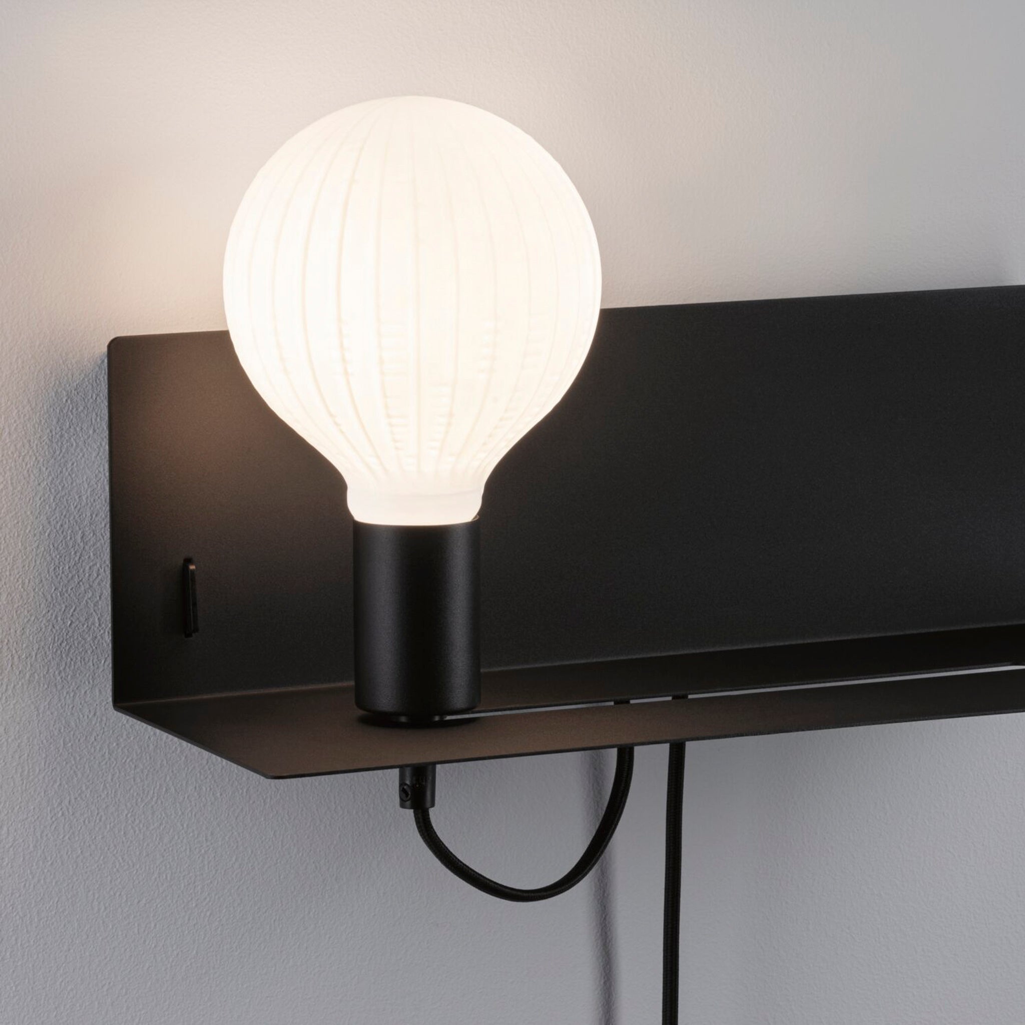 A modern Devara 40W Wall Mounted Lamp with USB Port in Black Matt features a white, glowing bulb on a sleek rectangular base and an integrated shelf, set against a light gray wall.