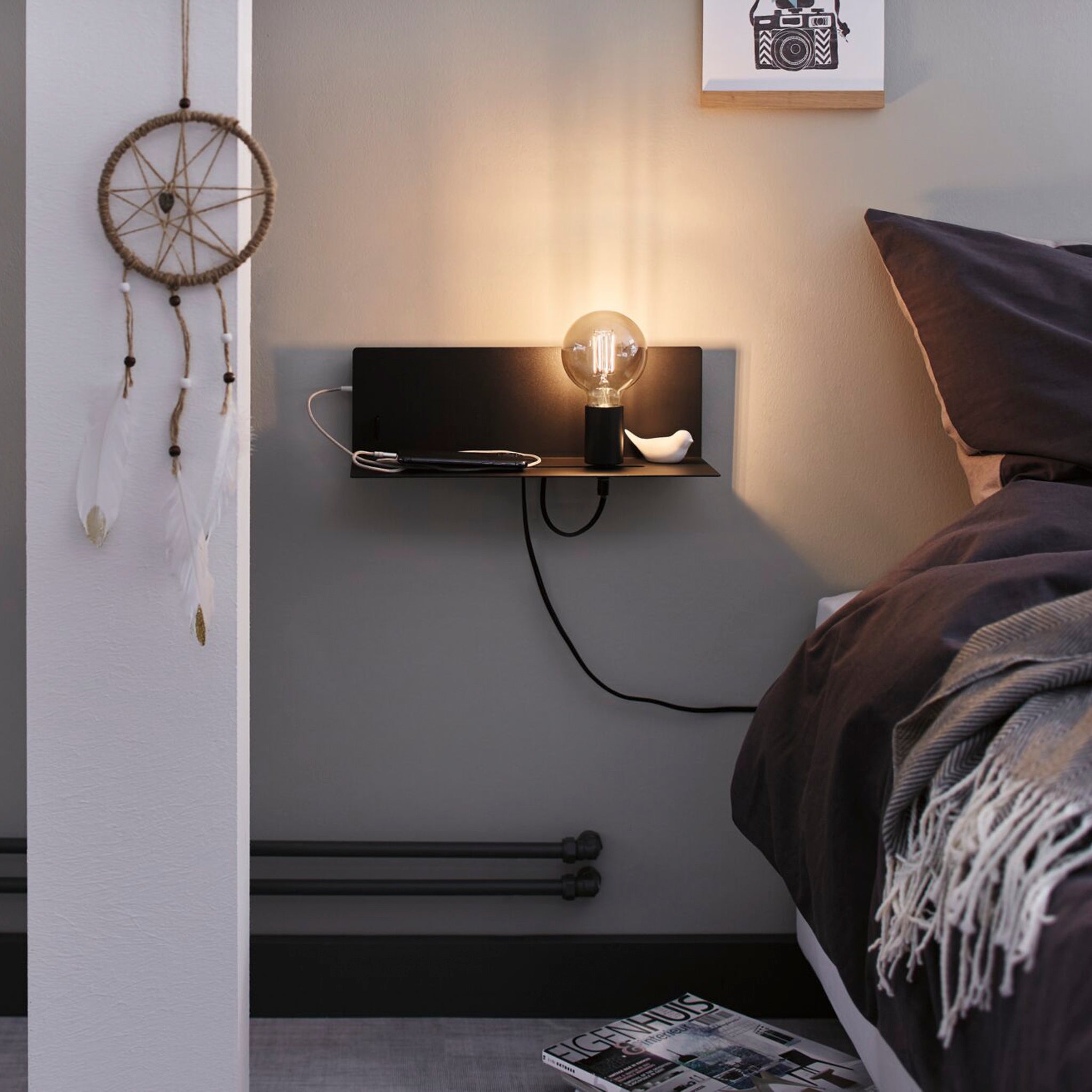 A modern bedroom features an integrated shelf holding a lit bulb and a small decor item. A dreamcatcher hangs on a nearby wall, adding charm, while the Devara 40W Wall Mounted Lamp with USB Port in Black Matt provides functional elegance.