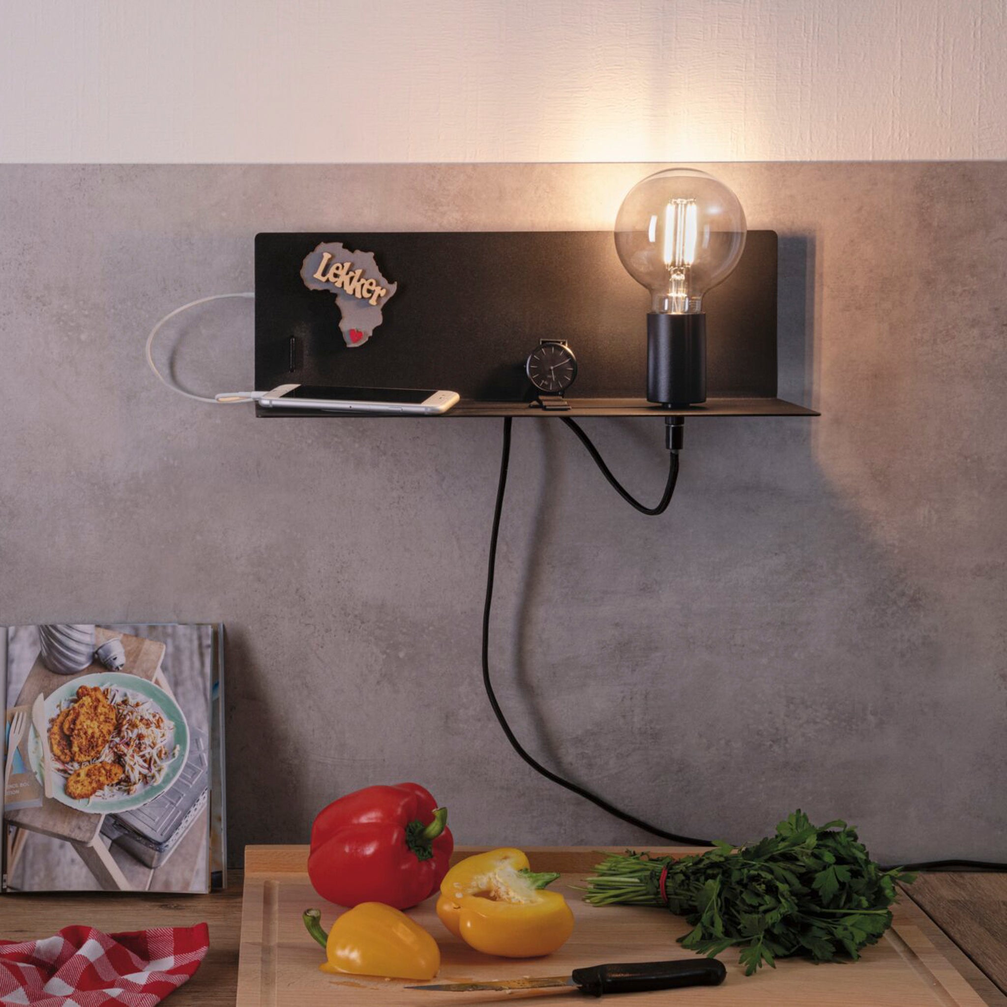 The Devara 40W Wall Mounted Lamp with USB Port in Black Matt, positioned over a kitchen counter adorned with vegetables, a knife, and a cookbook, also comes equipped with an integrated shelf and a convenient USB-C charging port.