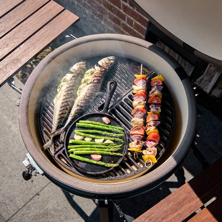 A Kamado Half Moon Cast Iron Griddle for 21" & 22" Grill, featuring the versatile Divide and Conquer Cooking System, showcases fish, vegetable skewers, and asparagus being cooked on a reversible griddle outdoors.