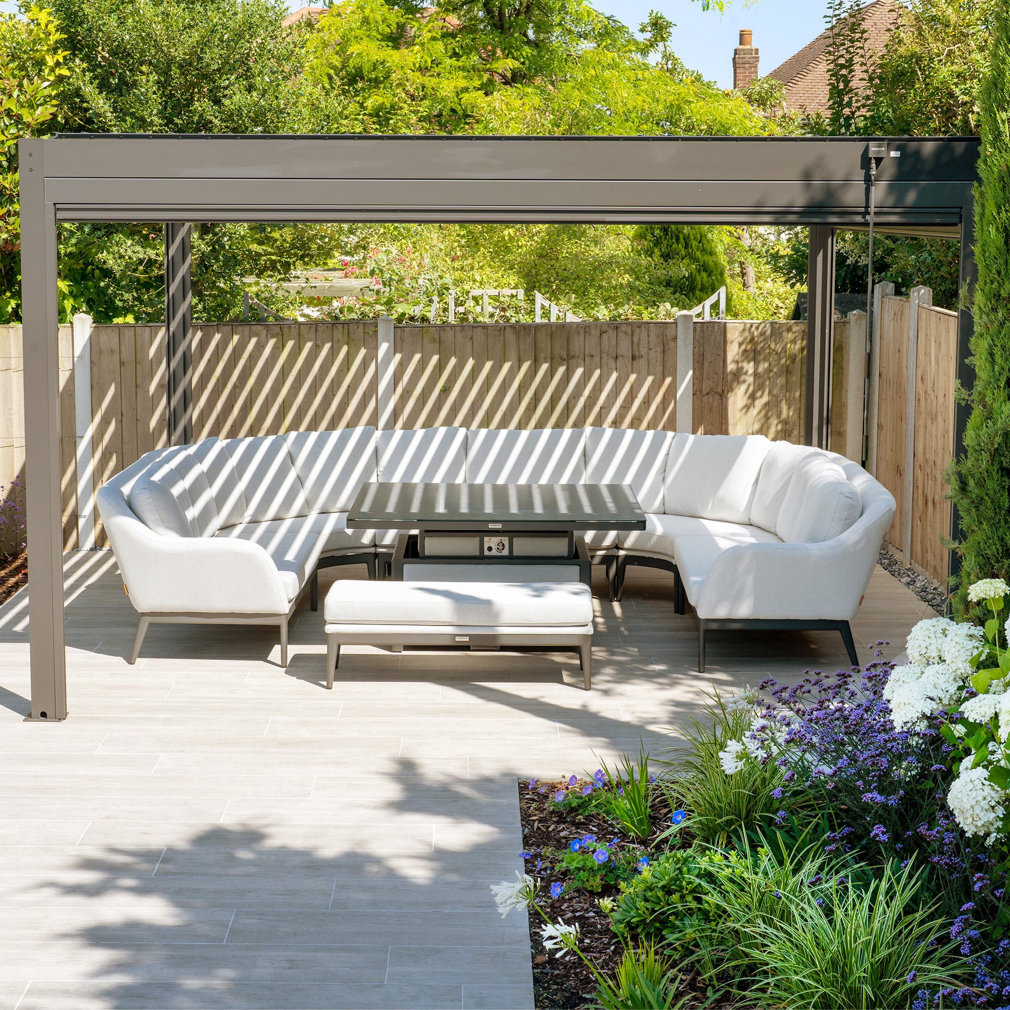 Modern outdoor patio featuring the Ali testing vimeo - no video Sofa Set with plush reflex foam cushions, a rectangular table under a sleek gray pergola, and a garden adorned with flowers. The sofa's hydrophobic thin rope weave adds a contemporary touch to the tranquil space.