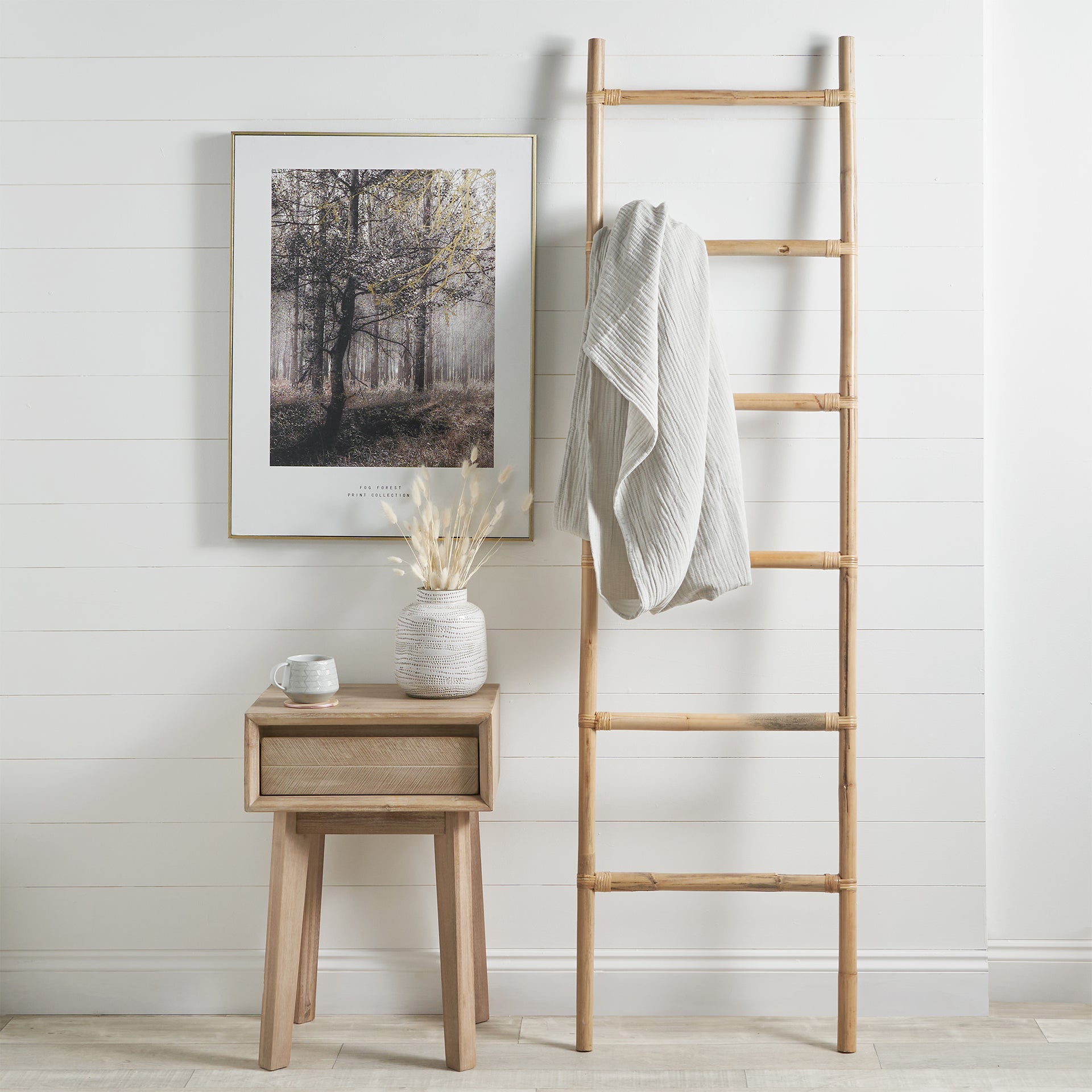 A wooden ladder with a draped blanket, a framed forest photo, and a small table showcasing the Alina Dot Design Small Stoneware Vase in White and a cup create a stylish scene.