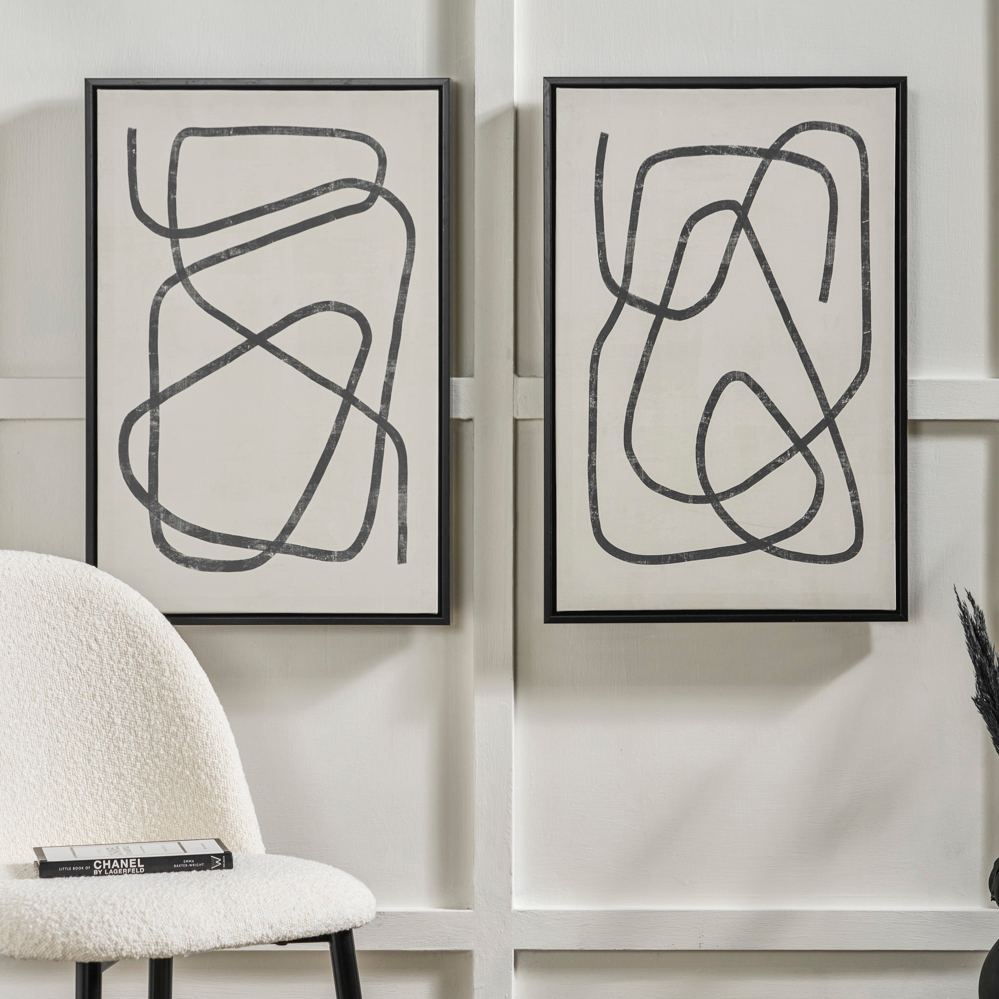 The Black Squiggle Print Canvases with Black Frames elegantly adorn a light wall, perfectly complementing a textured chair that holds several books on its seat, crafting a remarkable interior decor statement.