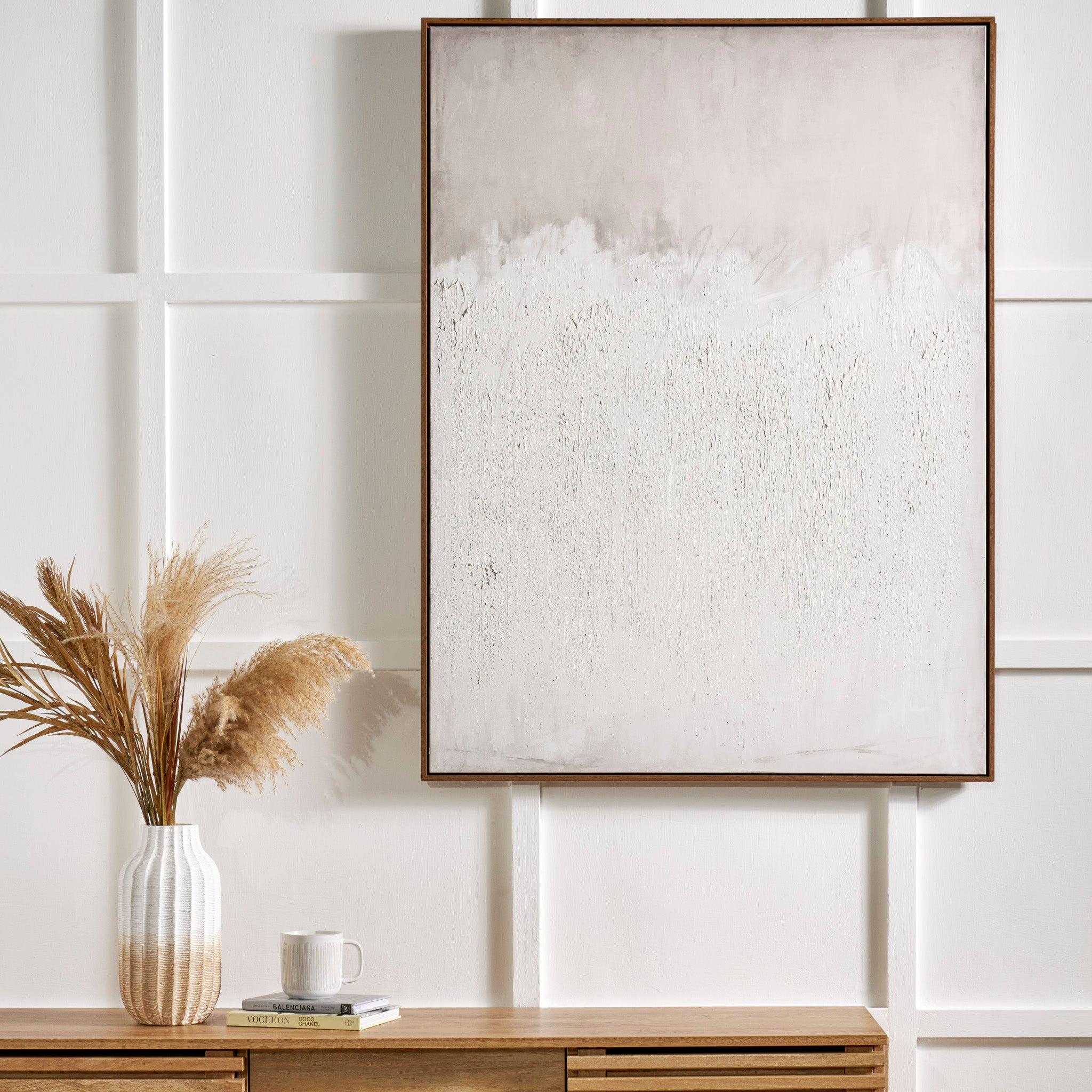 The Natural and White Abstract Canvas with a natural frame large size beautifully enhances the minimalist wall art above the wooden console, creating an inviting scene alongside a vase of dried pampas grass, a book, and a mug that complete this serene interior style.