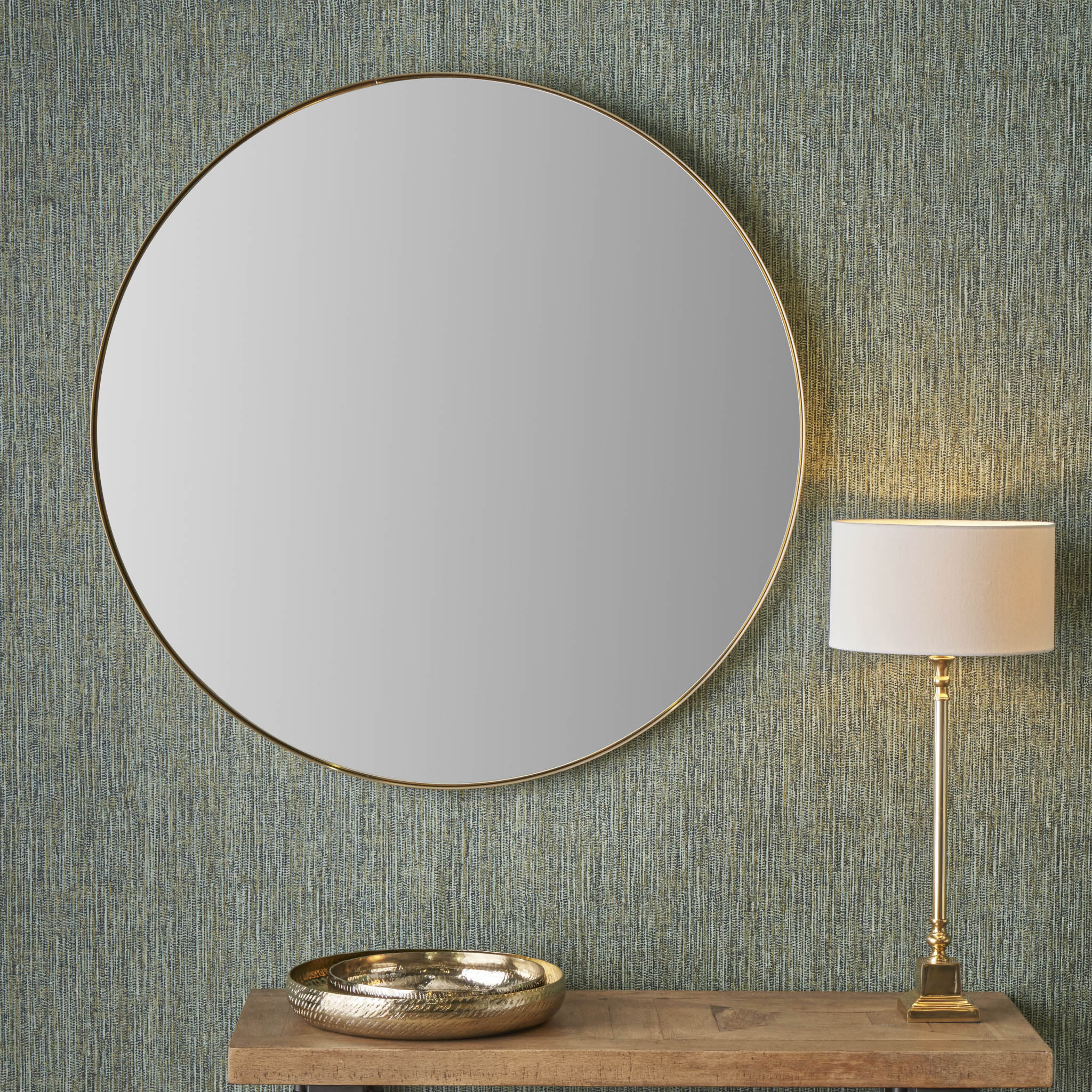 A Brushed Gold Metal Slim Frame Round Wall Mirror Large and a lamp rest on a wooden table against a textured green wall, showcasing minimalist design with a touch of elegance. The lamp's brushed gold finish beautifully complements the mirror.