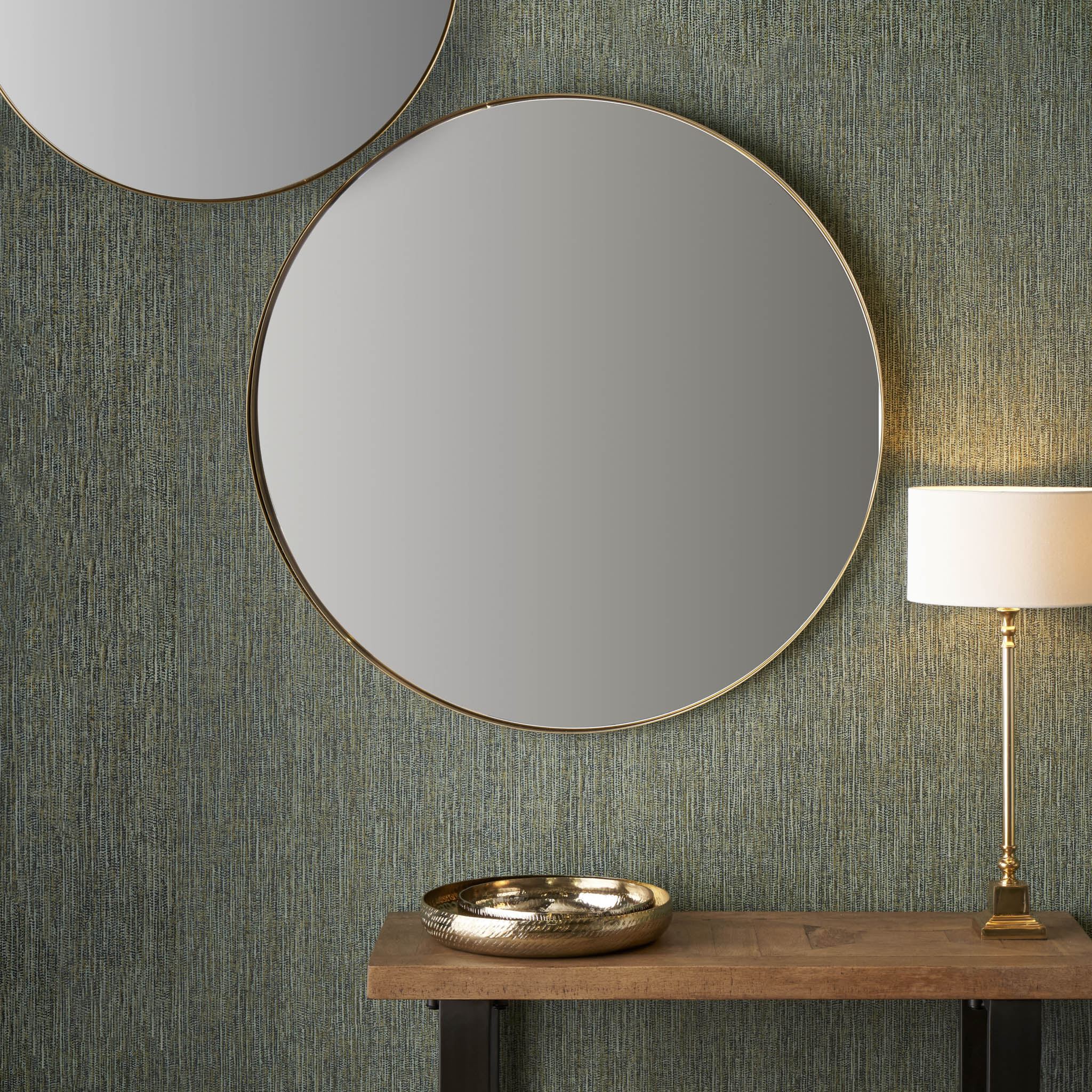 A contemporary arrangement features the Brushed Gold Metal Slim Frame Round Wall Mirror Large and a lamp on a wooden table, accompanied by a decorative bowl, all set against a textured green wall. The minimalist design is enhanced by the brushed gold finish of the mirror's frame, adding an elegant touch.